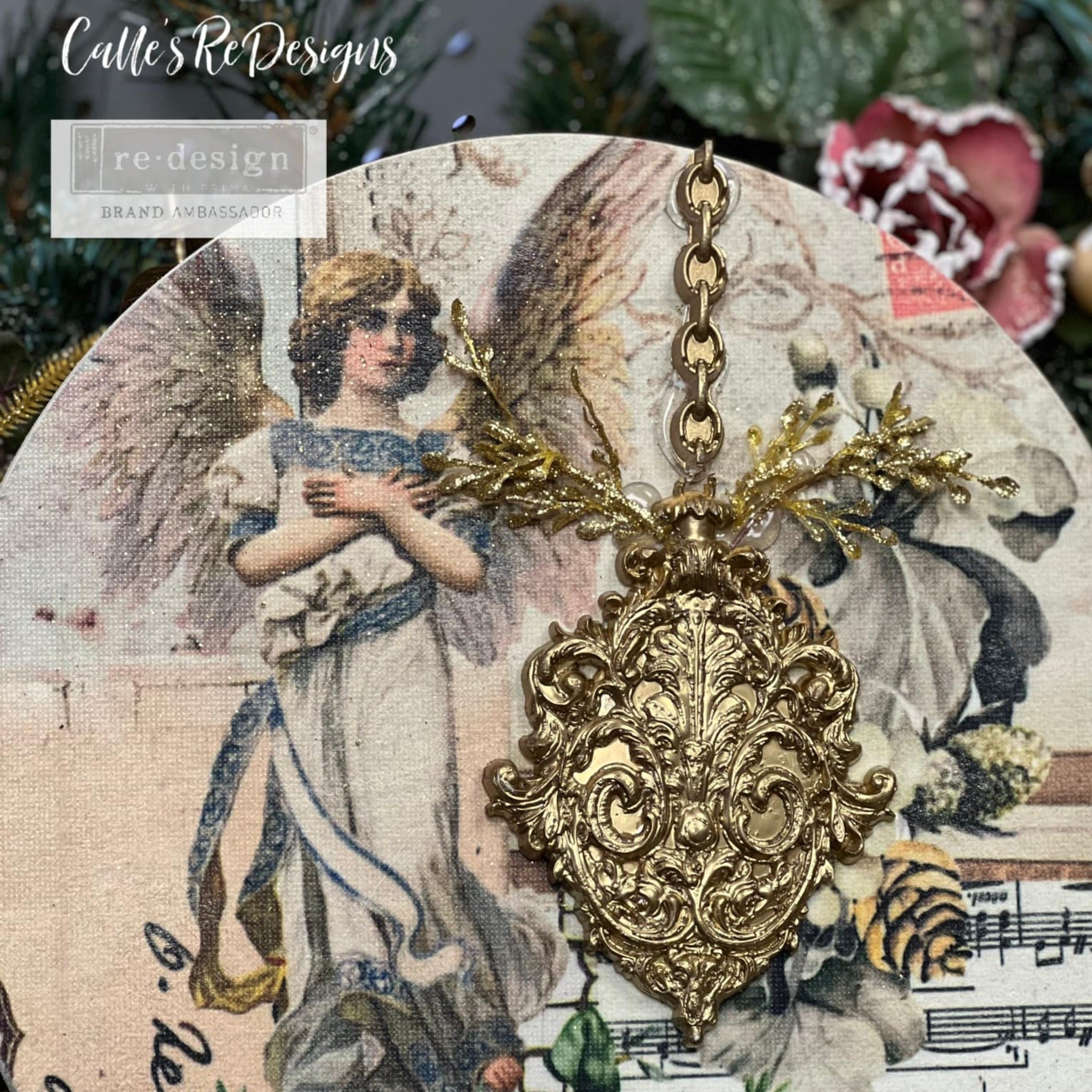 A circle canvas created by Calle's ReDesigns features an angel and vintage sheet music from ReDesign with Prima's Holly Jolly Hideaway 3 pack tissue papers. A gold painted silicone mould casting of an ornate ornament is also adorning the canvas.