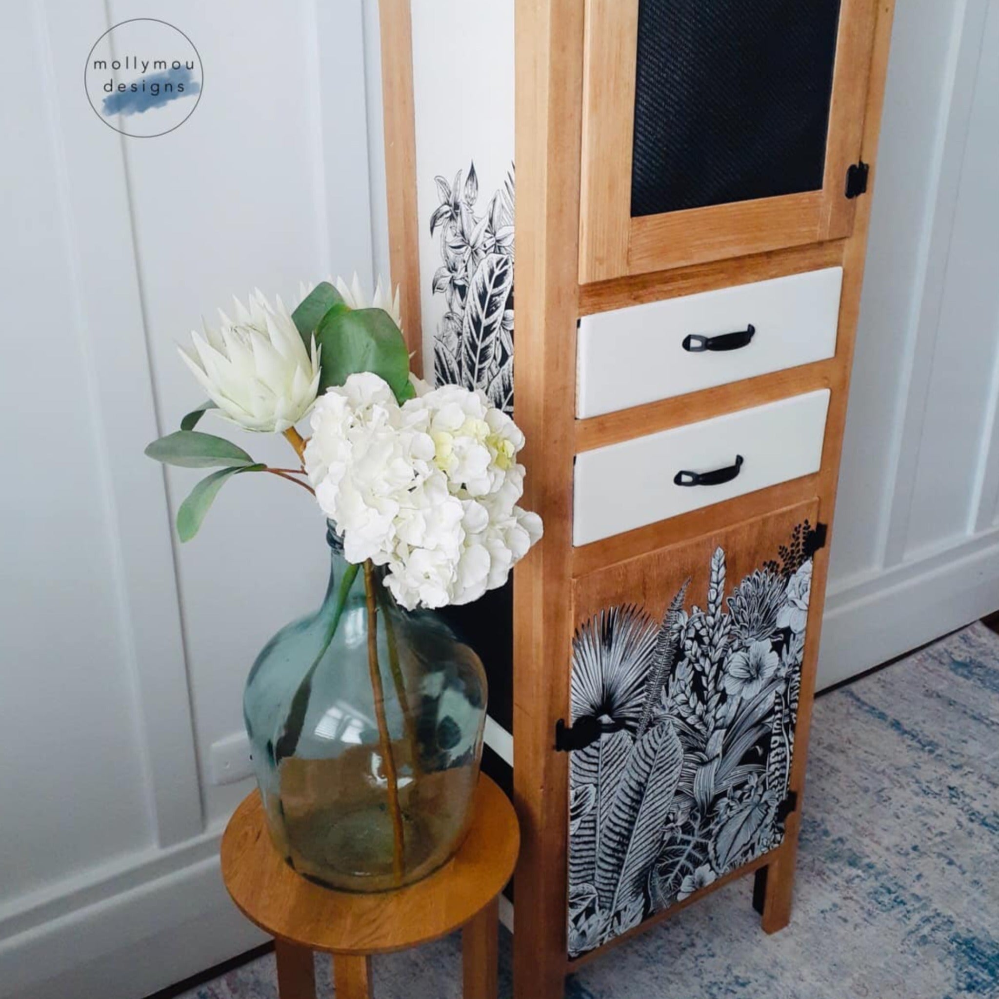 A tall, narrow storage hutch refurbished by Mollymou Designs is natural wood with white drawers and white side panels and features ReDesign with Prima's Abstract Jungle transfer on the white side panel and on a natural wood bottom door.