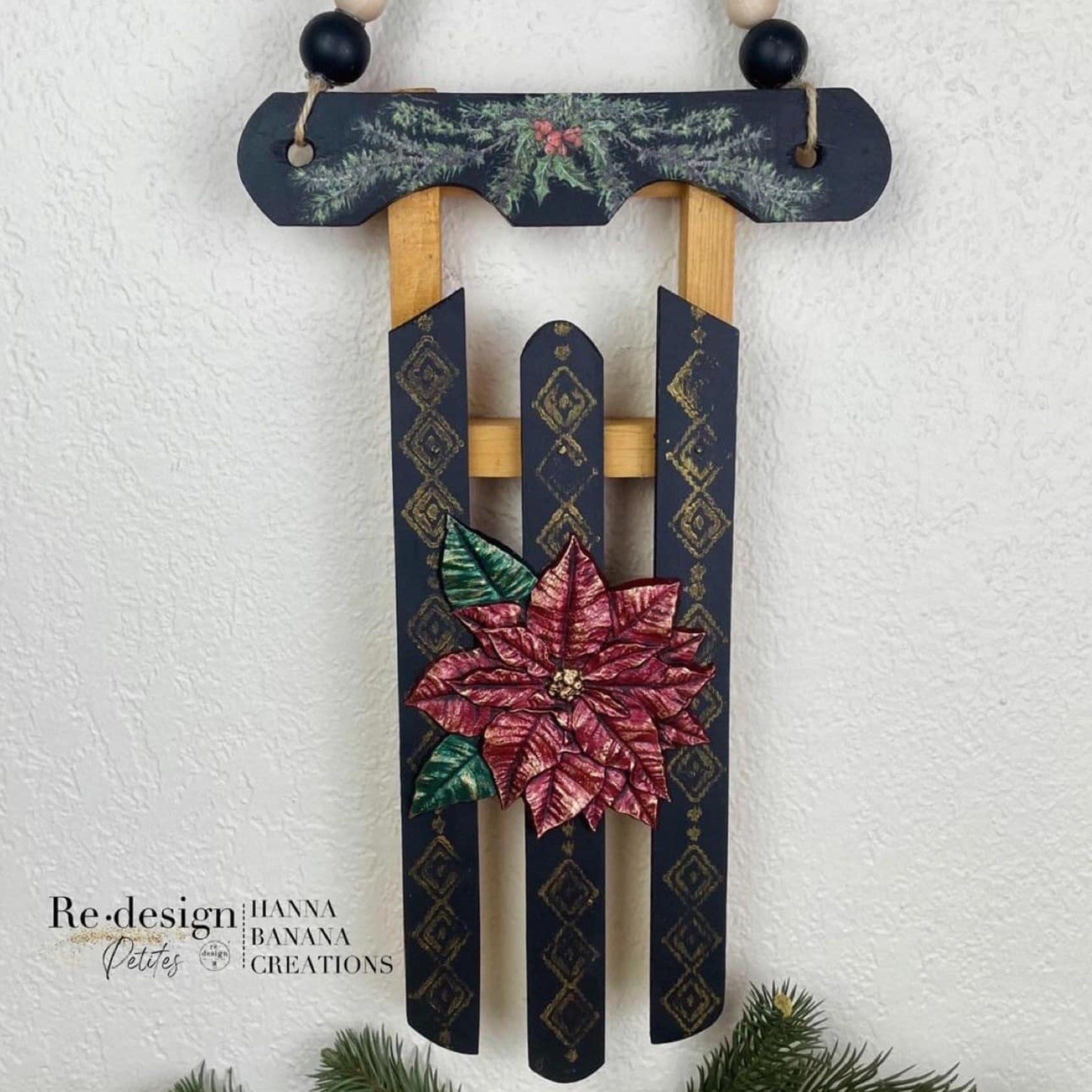A sleigh decoration refurbished by Hanna Banana Creations is painted navy blue with natural wood and features ReDesign with Prima's Perfect Poinsettia silicone mould casting in metallic red and metallic green on the center of the seat.