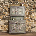A vintage hutch-top console table against a rustic stone backdrop is painted a distressed blend of grey, green, and brown and features ReDesign with Prima's Blossom Botanica transfer on it.