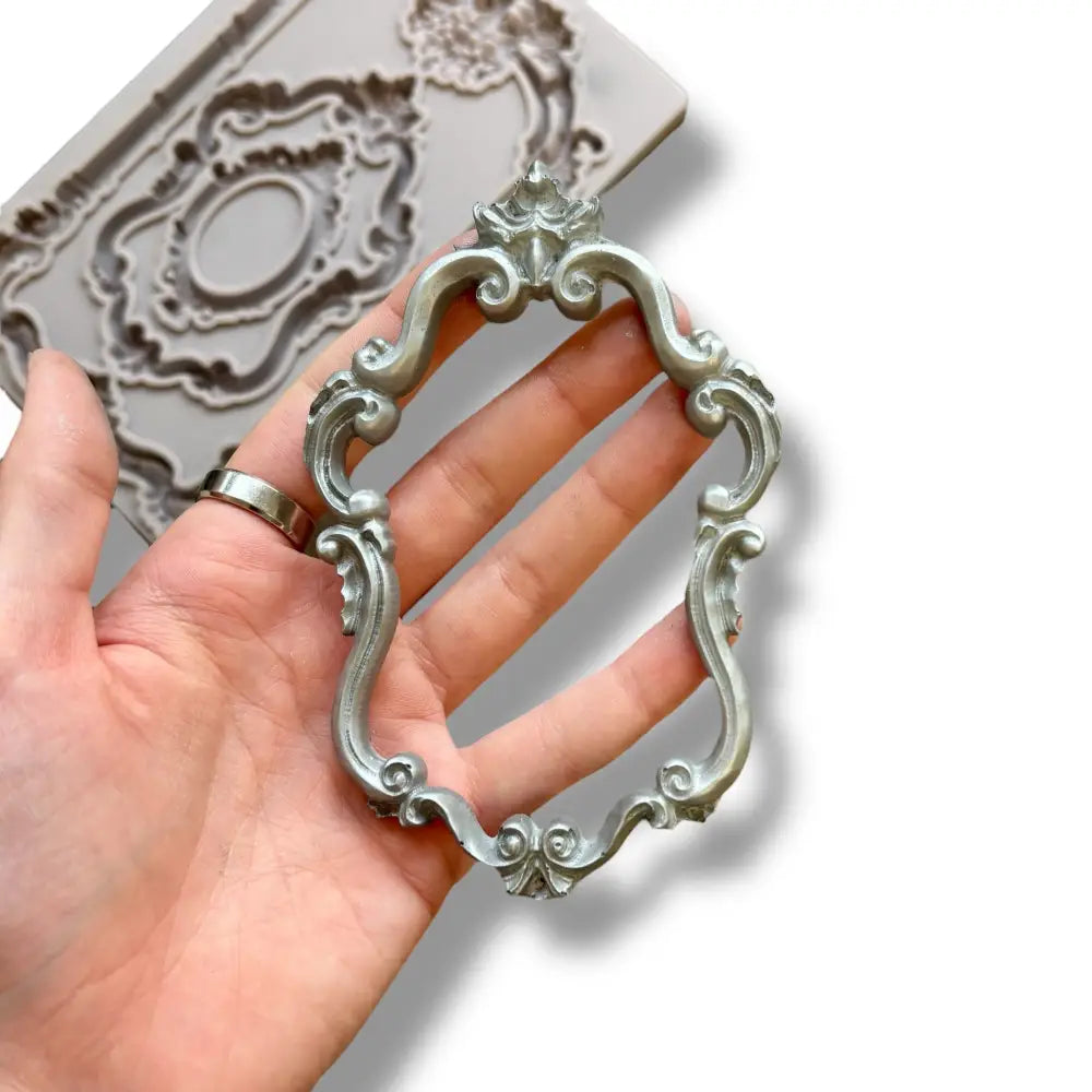 A light grey silicone mold featuring 3 intricately detailed baroque flourish frames is against a white background. A hand is shown holding a silver colored casting of one of the frames.