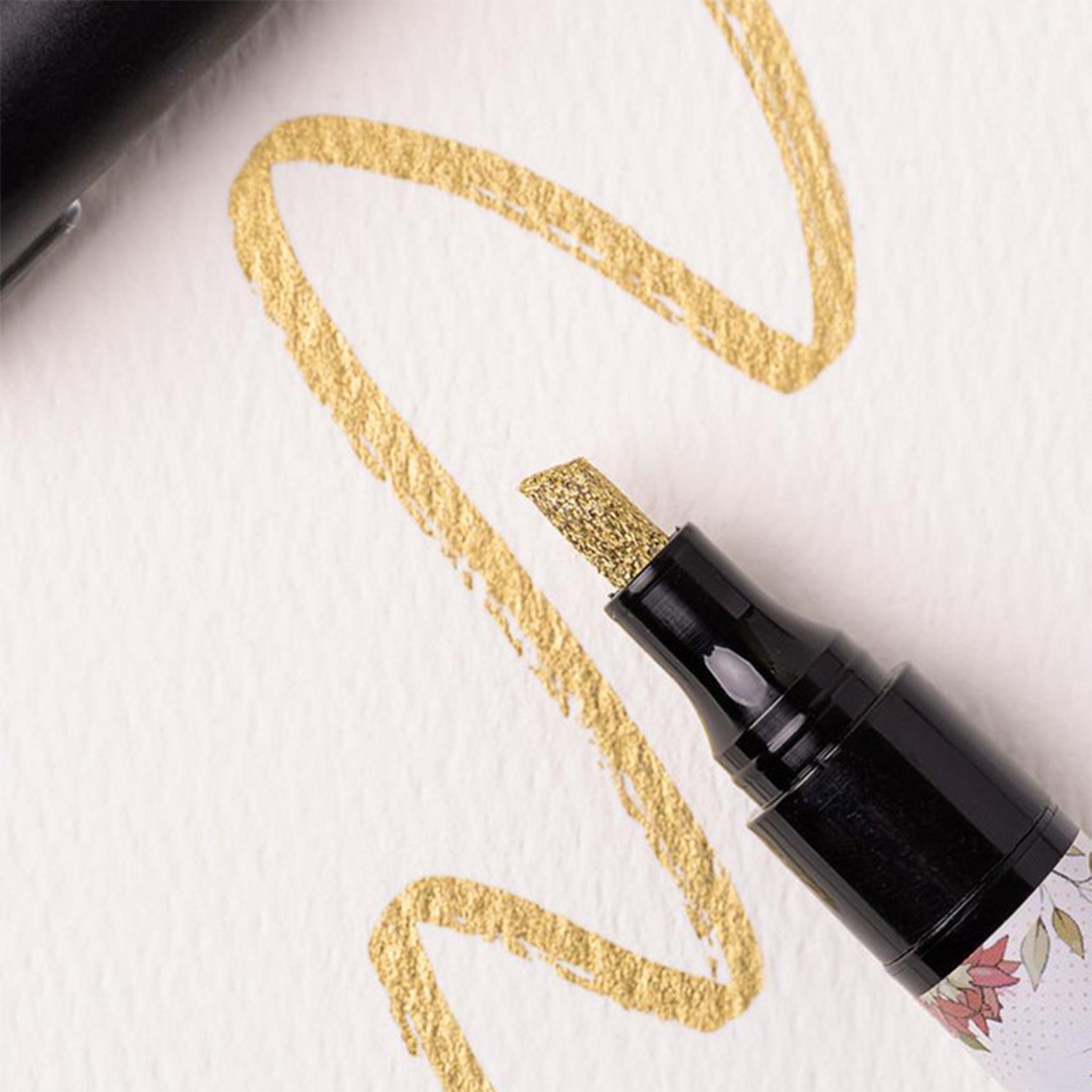 A close-up of Belles and Whistles' Gold Metallic Paint Pen chisel tip is being shown on white cardstock with a scribble example of the pen.