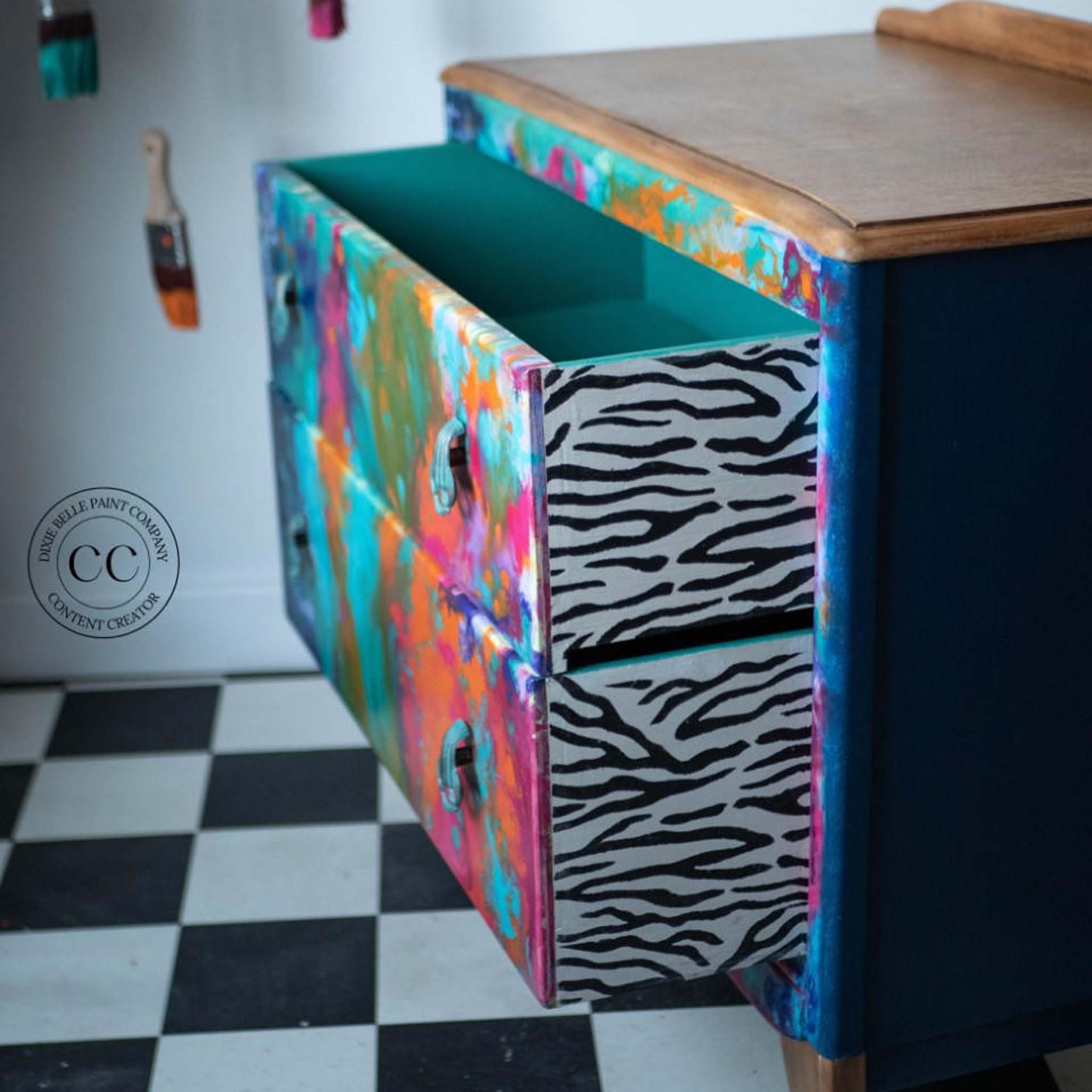 A 2-drawer nightstand is painted blue on the sides, natural wood on top, and artsy colorful paint splatter on the front. The sides of the drawers feature Belles & Whistles Safari mylar stencil zebra pattern in black and white.