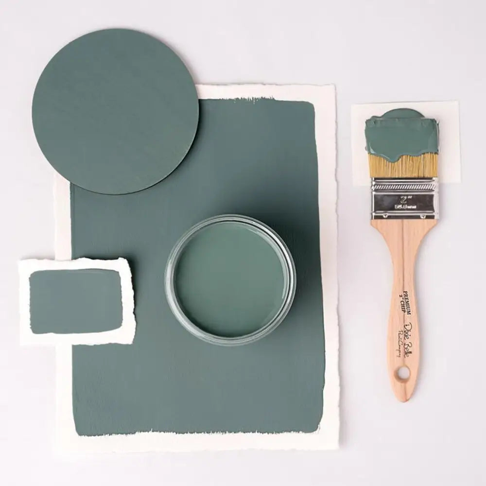 A paint brush, paper, and round wood samples of Dixie Belle's Blue Grass Chalk Mineral Paint are against a white background.