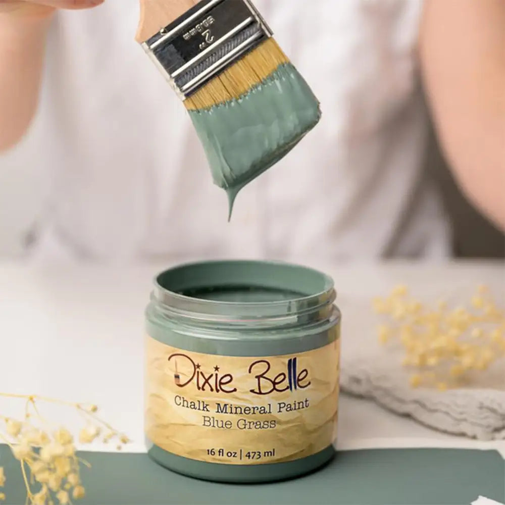 A paint brush with dripping paint is shown above an open container of Dixie Belle's Blue Grass Chalk Mineral Paint.