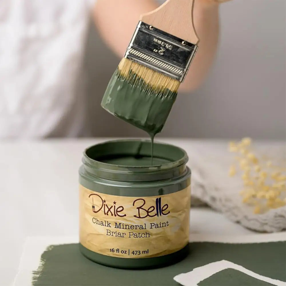 A paint brush with dripping paint is shown above an open container of Dixie Belle's Briar Patch Chalk Mineral Paint.