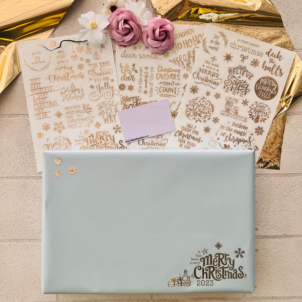 A light blue paper wrapped present and three sheets of Get Inspired's Christmas Greetings Gold Foil small transfers are against a textured beige backgroundwith sheets of gold foil near the top.