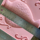 Pink decorative silicone mold with intricate patterns and a ruler for scale on a wooden surface.