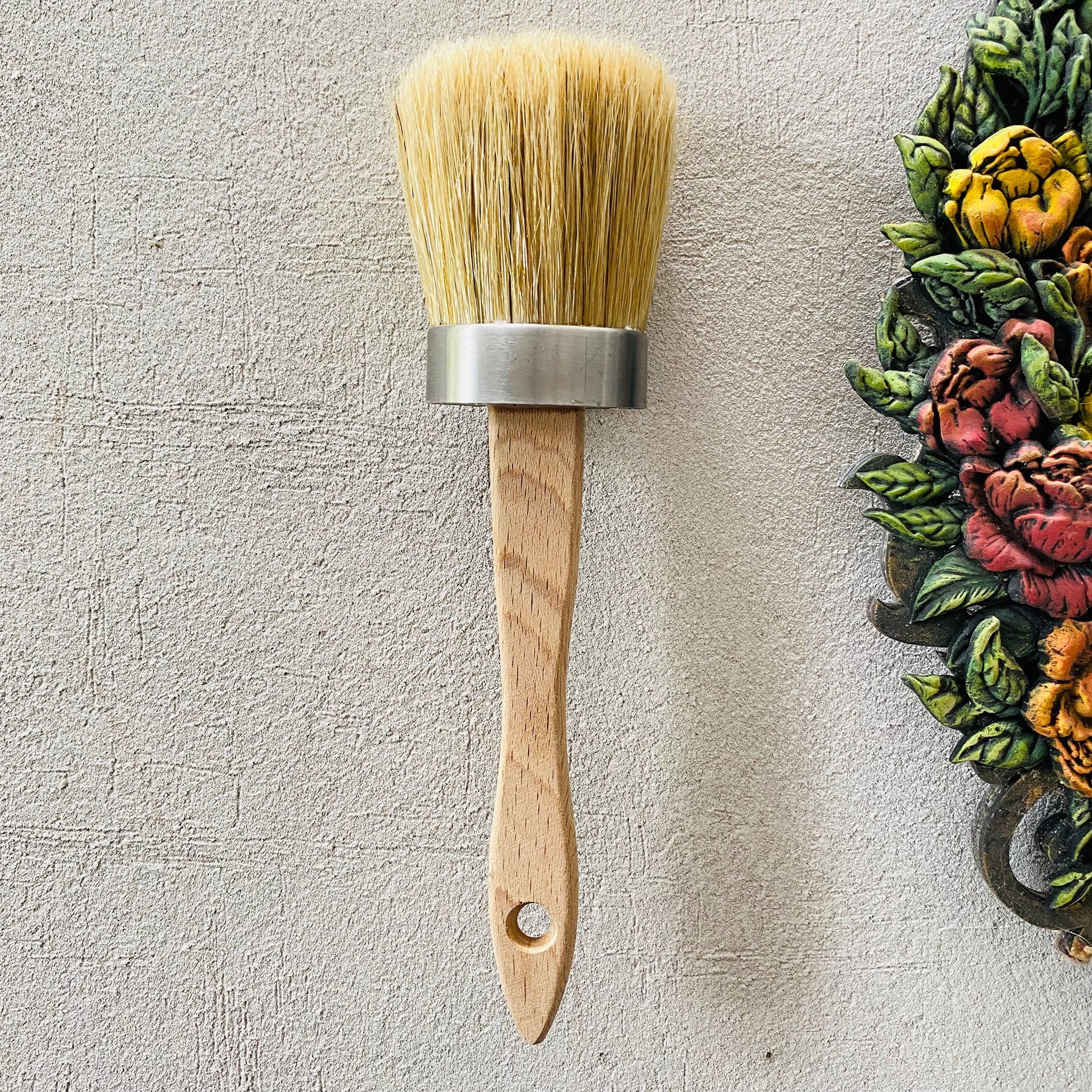 An oval bristle paint brush with a wooden handle by Dadarkar Arts by Get Inspired is against a textured light grey background. Part of a colorful floral silicone mold casting is to the right of the brush.