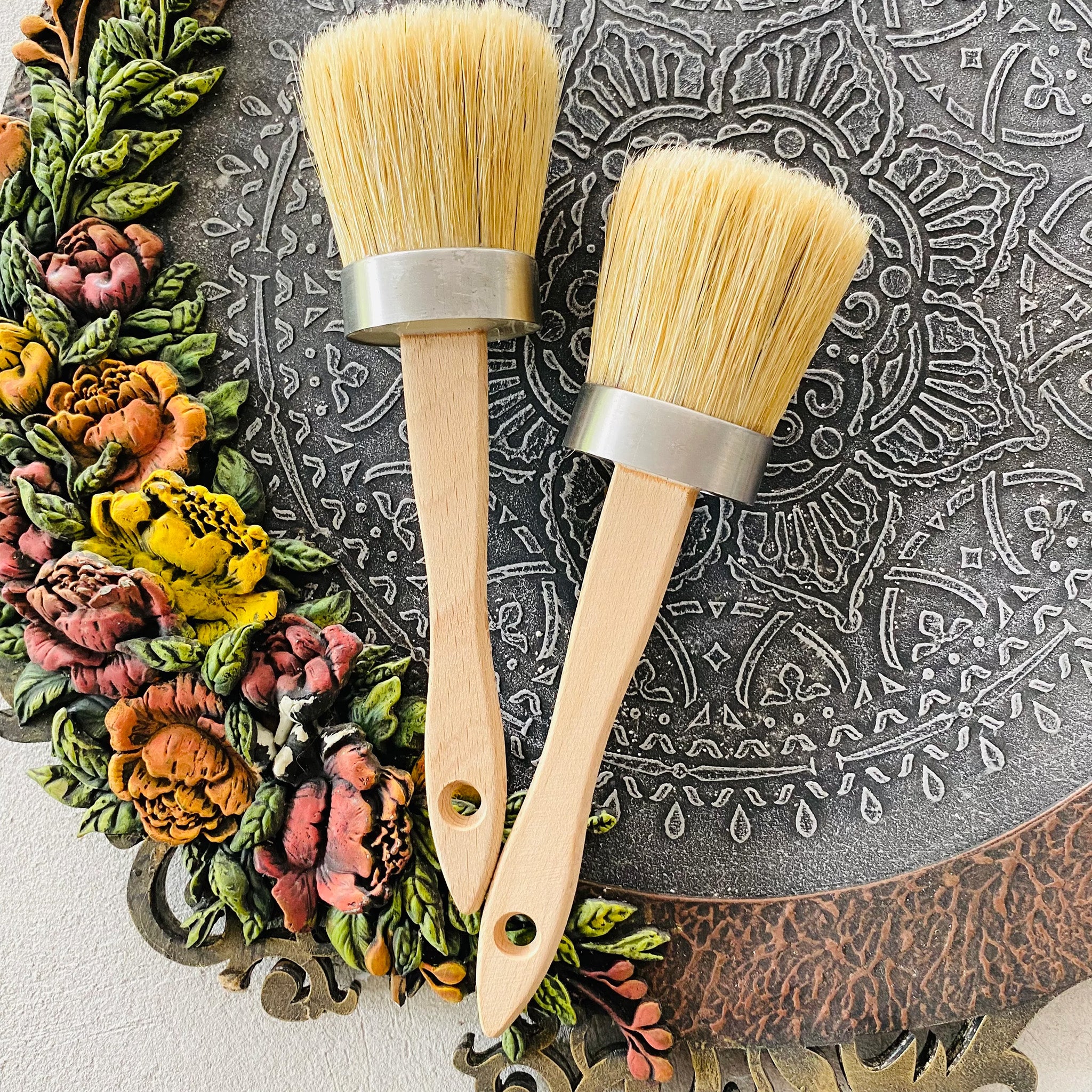 Two oval bristle paint brushes with wooden handles are shown against a stone mandala decor.