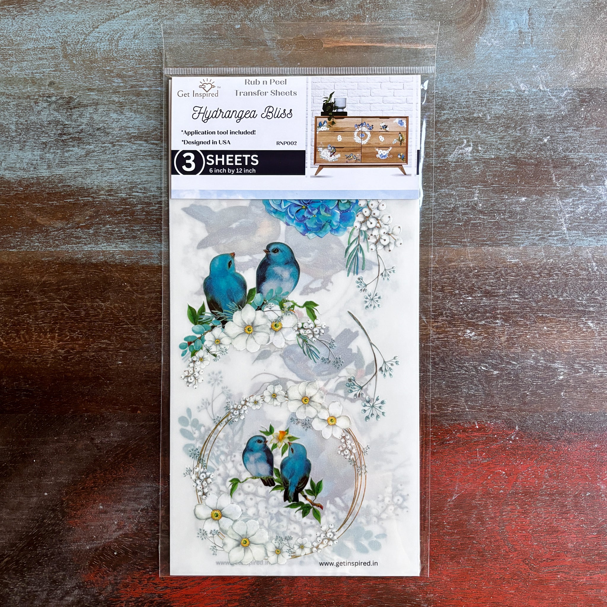 A package of Dadarkar Art’s Get Inspired Hydrangea Bliss rub-on transfers is against a dark wood background.