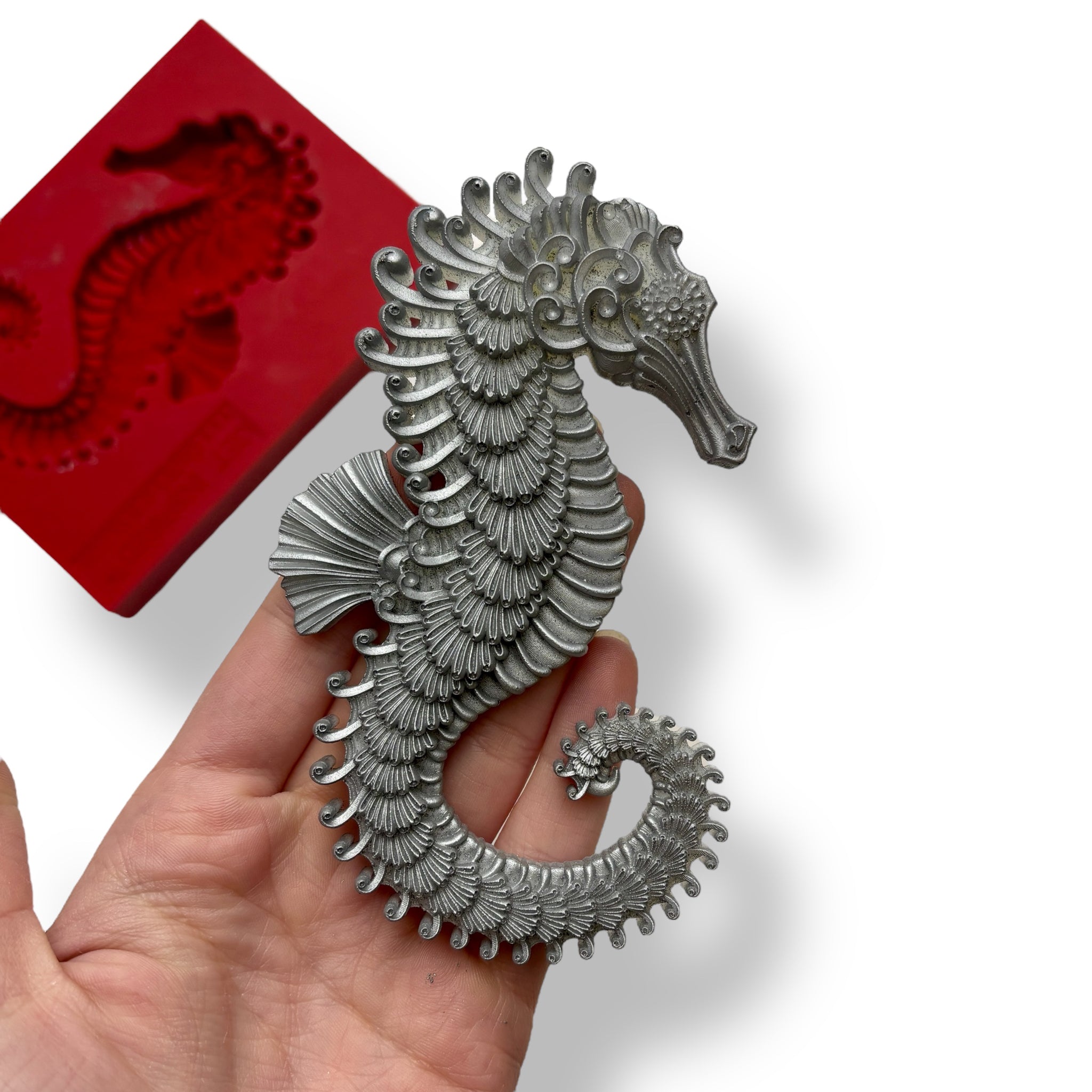 A red silicone mold and silver colored casting of a seahorse are against a white background. A hand is shown holding the casting.