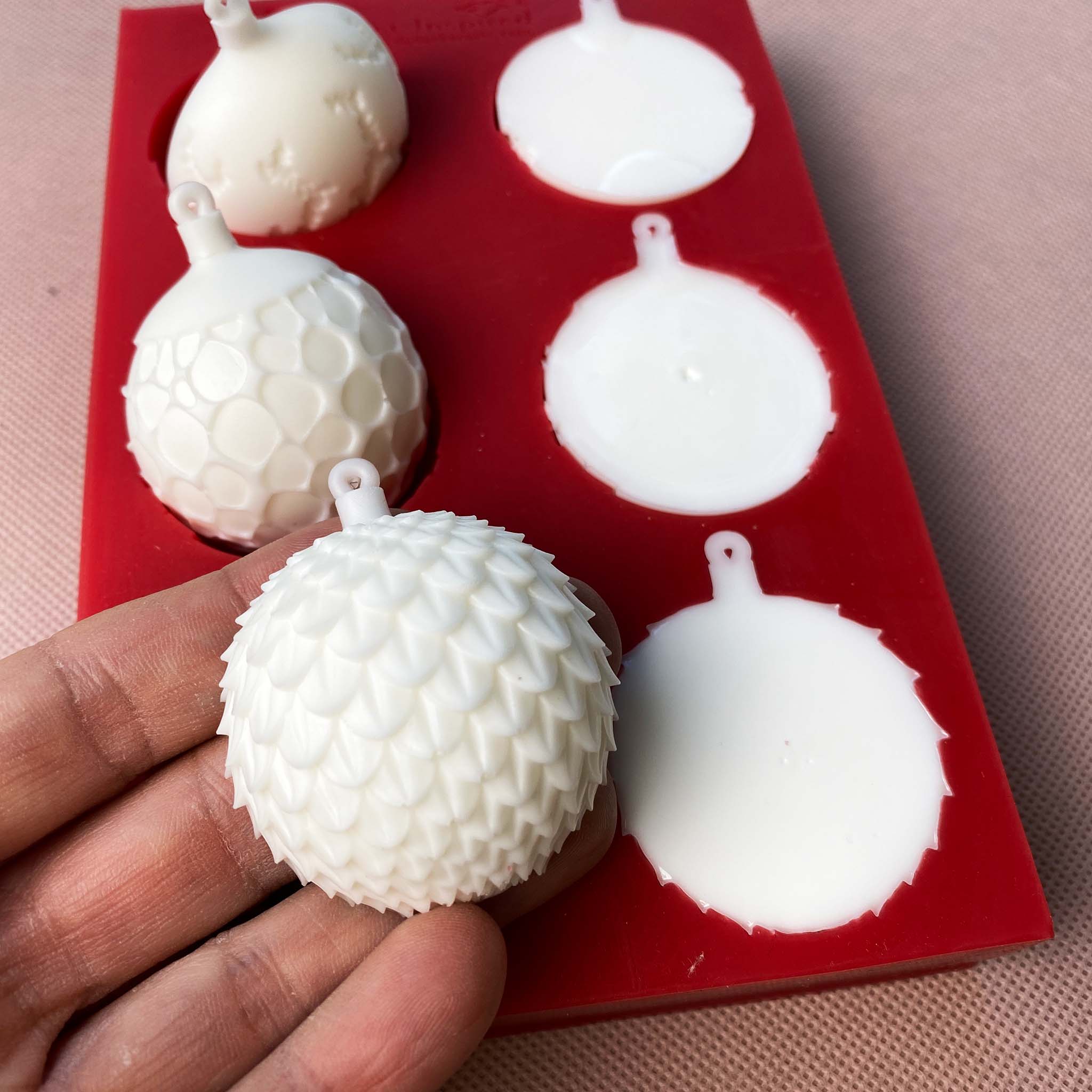 A red silicone mould of Get Inspired by Dadarkar Art's Christmas Ornaments is against a light colored material background. White resin castings sit in 3 of the moulds; 2 are flipped up to show the design; and a hand is holding one of the castings on 3 fingertips..