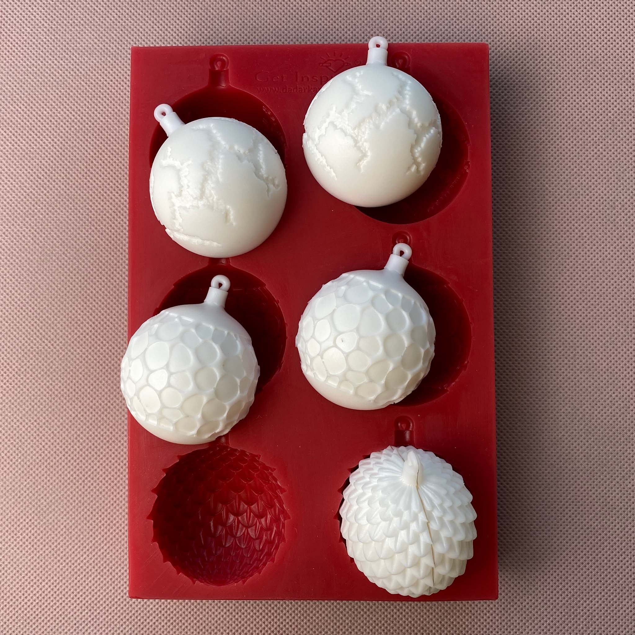 A red silicone mould of Get Inspired by Dadarkar Art's Christmas Ornaments is against a light material background. On the mould are white resin castings of the ornaments, the bottom of which shows how 2 halves can be put together to create a full ornament.