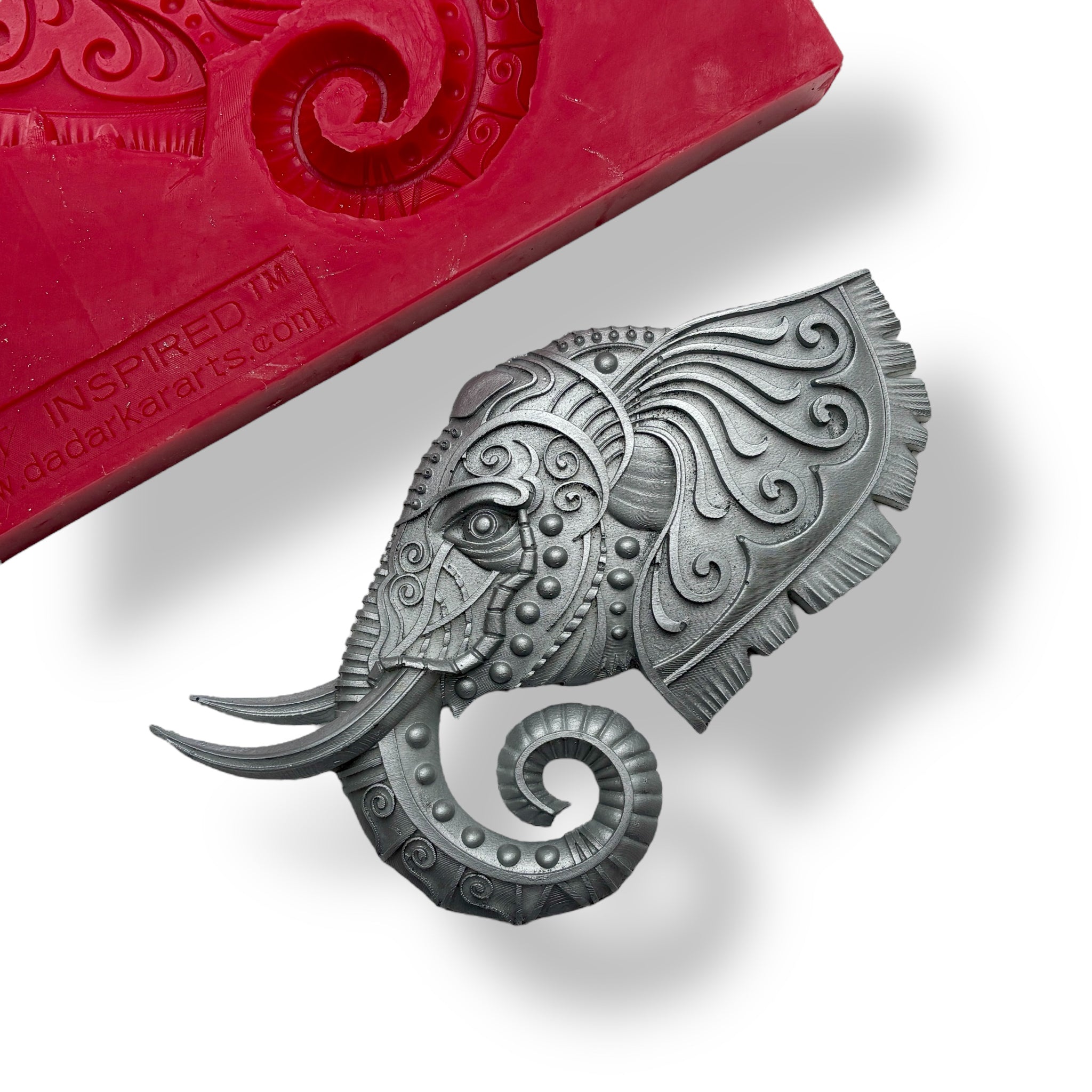 A red silicone mold and silver colored casting of an elephant head with ornate adornments are against a white background.