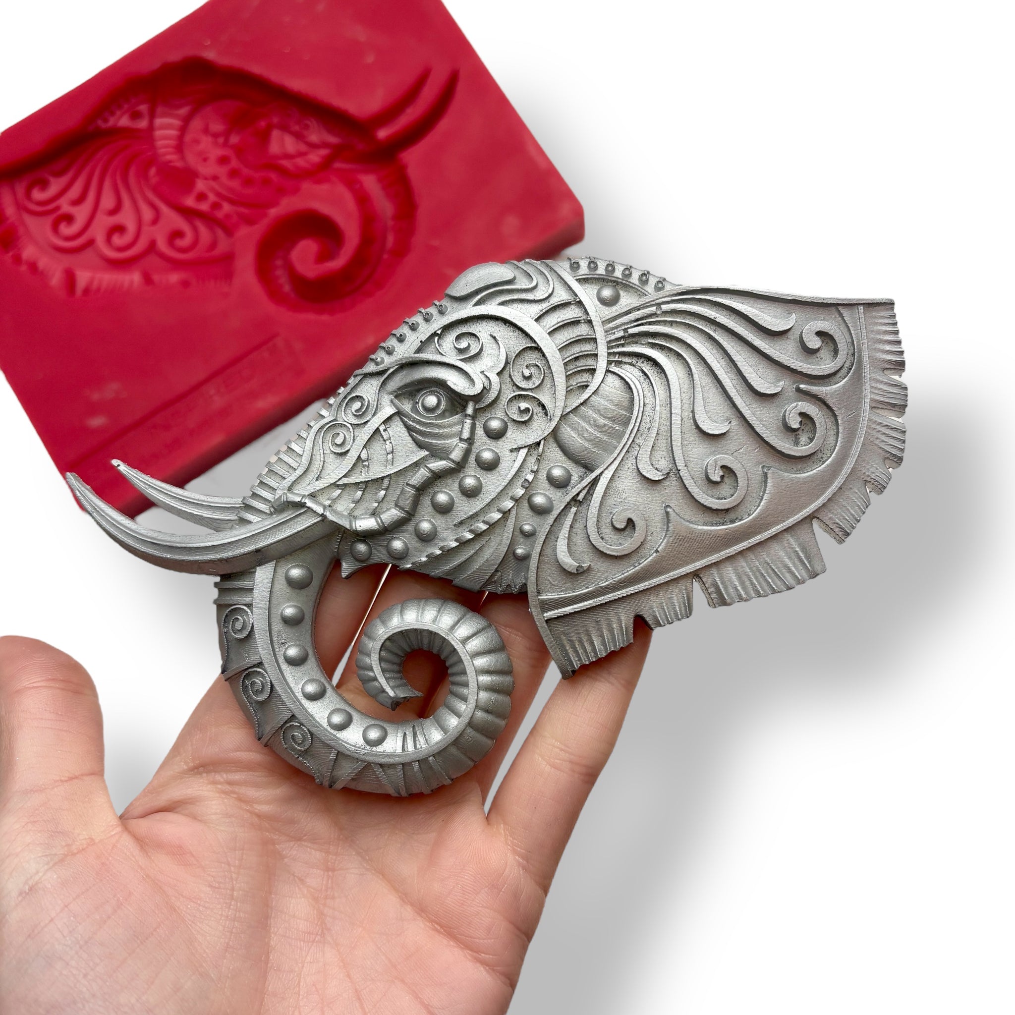 A red silicone mold and silver colored casting of an elephant head with ornate adornments are against a white background. A hand is shown holding the casting.