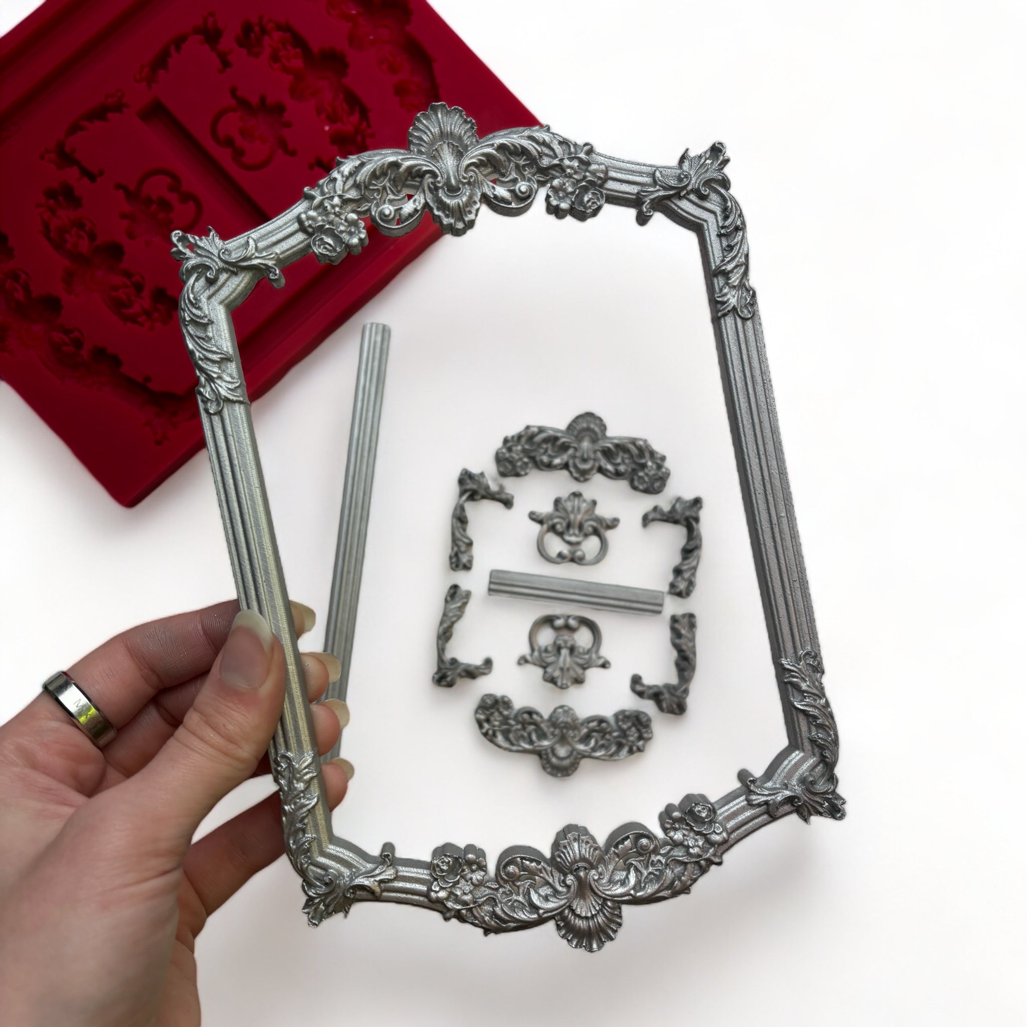 A red silicone mold and silver colored castings of ornate vintage frames and borders are against a white background. A hand is shown holding the large frame.