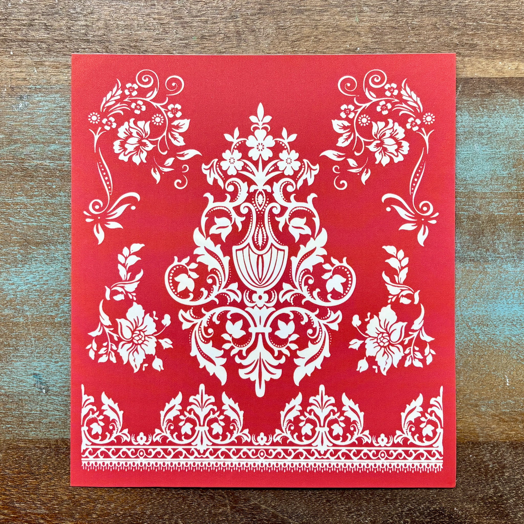 A red silkscreen stencil featuring an ornamental baroque pattern and floral flourishes is against a wood background.