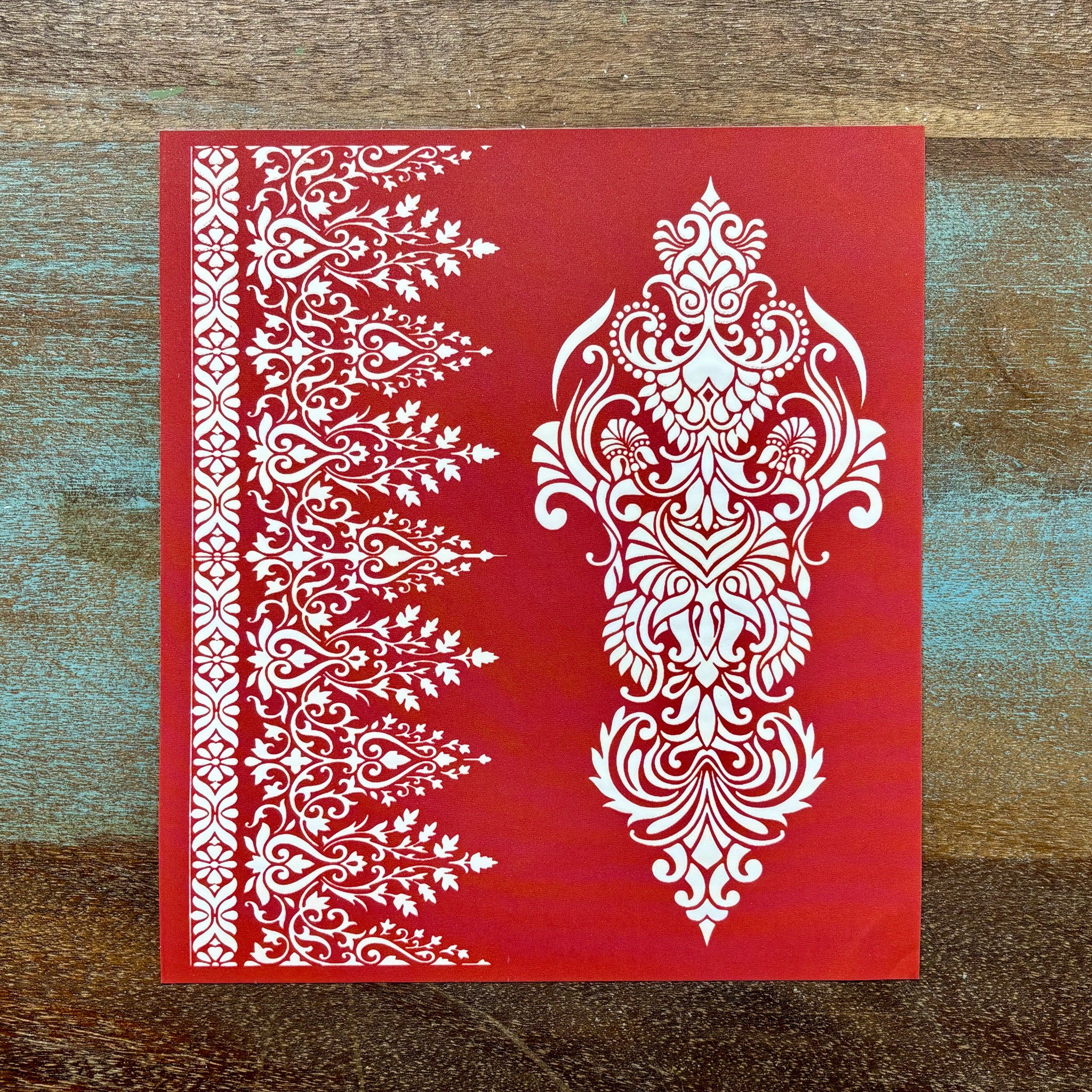 A red silkscreen stencil featuring a large paisley flourish and a smaller repeating paisley border is against a wood background.