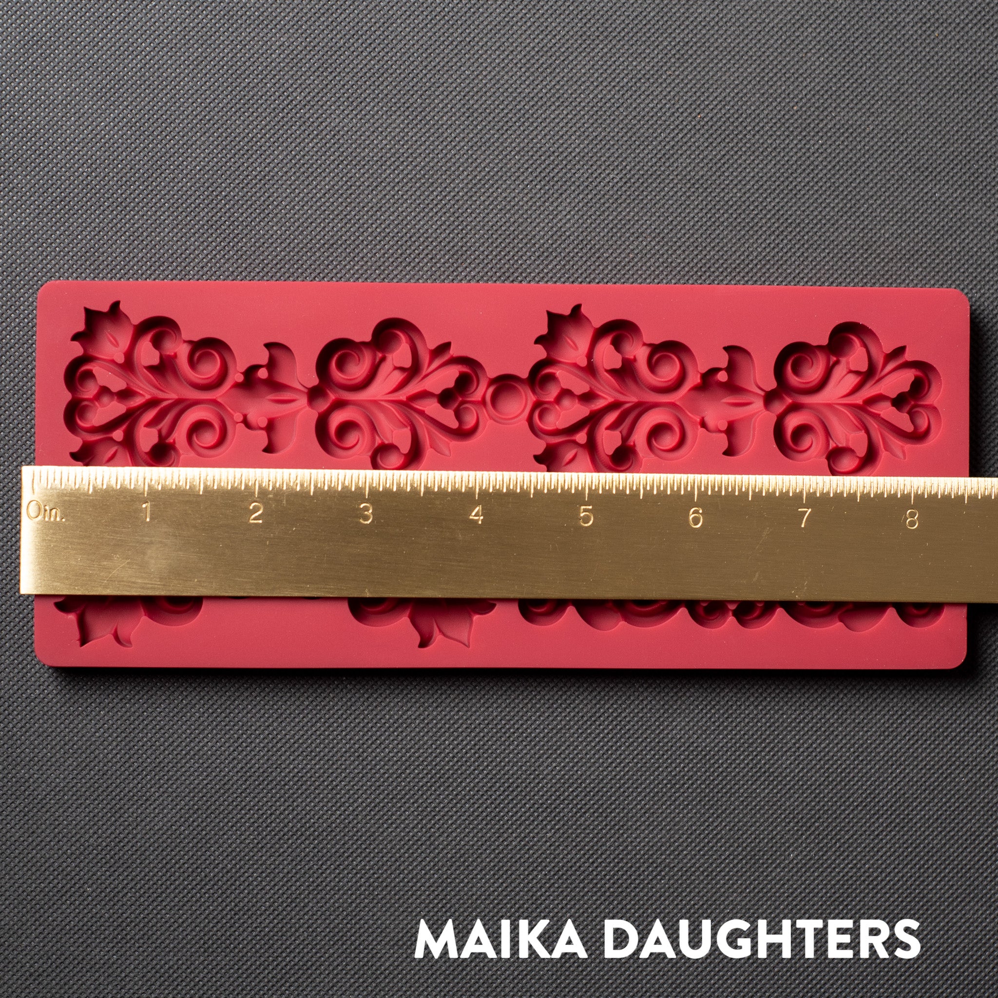 A red silicone mold that features 4 small corner flourishes and one larger border is against a dark grey material background. A gold ruler reading 8.5" length sits on the mold.