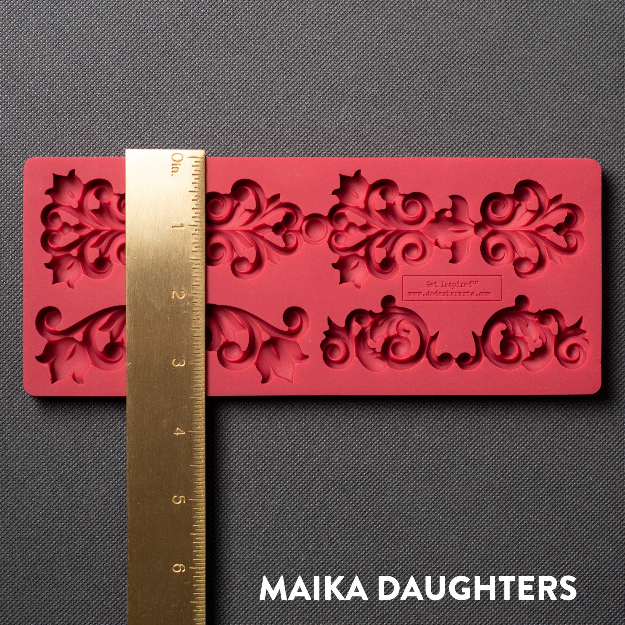 A red silicone mold that features 4 small corner flourishes and one larger border is against a dark grey material background. A gold ruler reading 3.5" height sits on the mold.