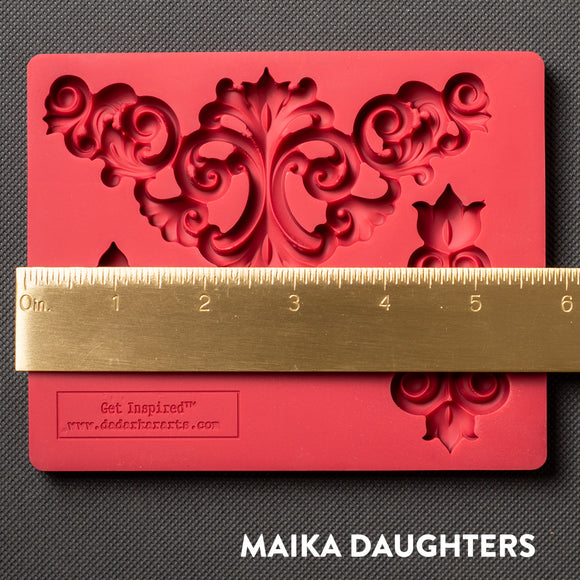 A red silicone mold that features 3 avant-garde style flourish corner pieces is against a dark grey material background. A gold ruler reading 5.75" length sits on the mold.