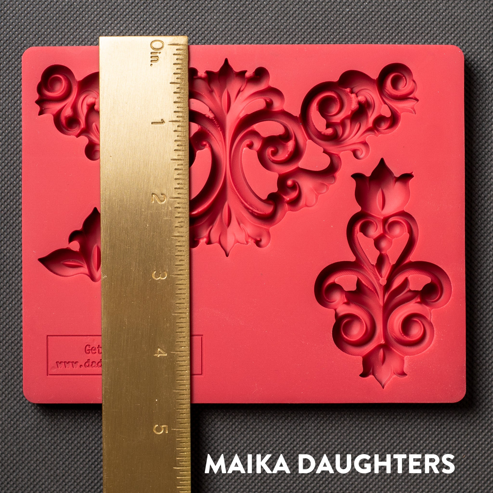 A red silicone mold that features 3 avant-garde style flourish corner pieces is against a dark grey material background. A gold ruler reading 4.5" height sits on the mold.