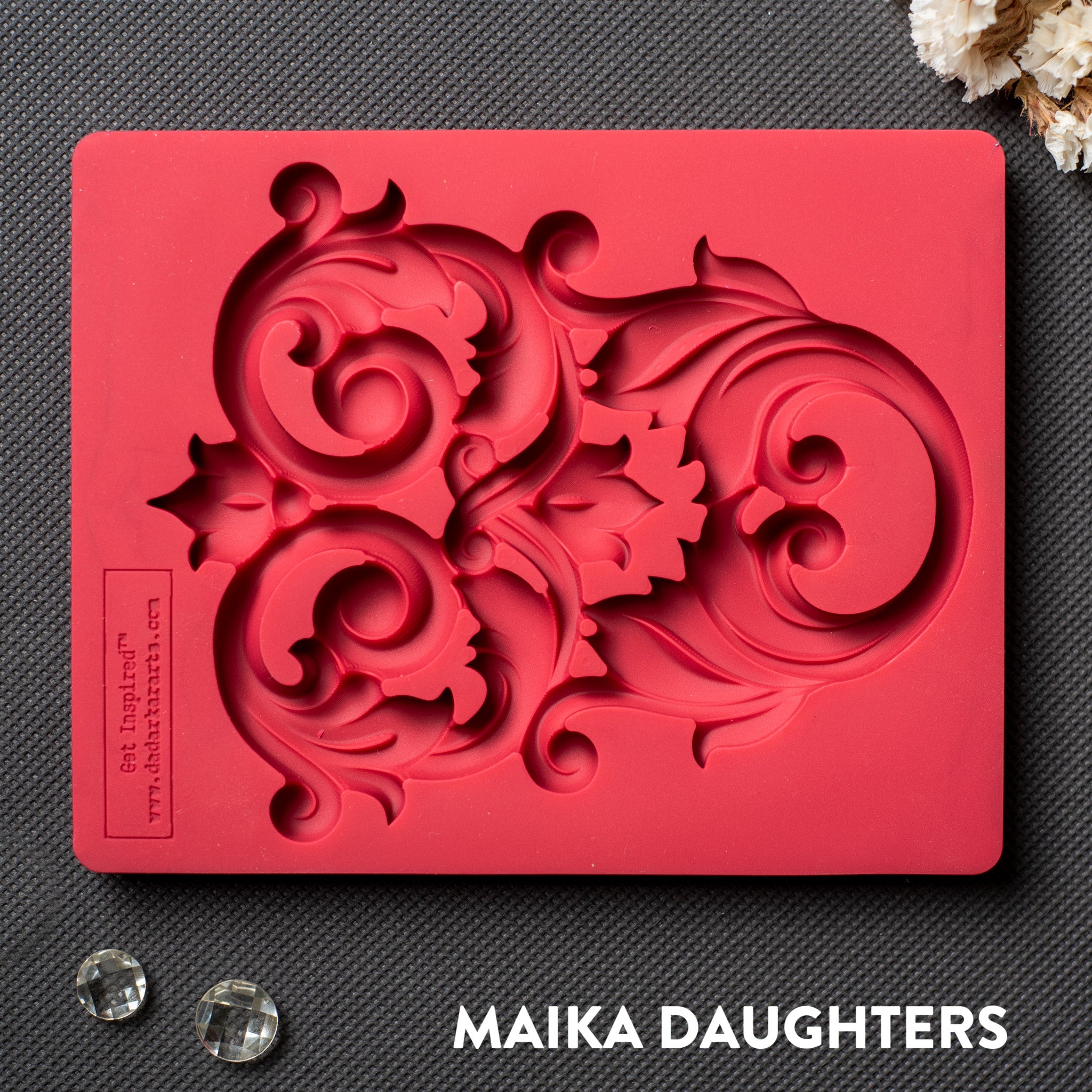 A red silicone mold that features an intricate flourish accent piece is against a dark grey material background.