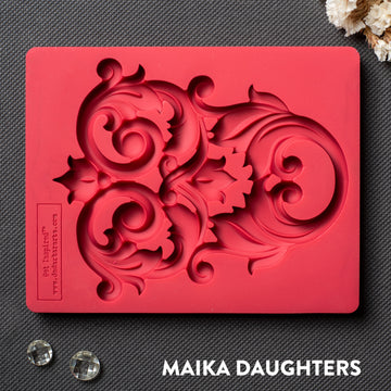 A red silicone mold that features an intricate flourish accent piece is against a dark grey material background.