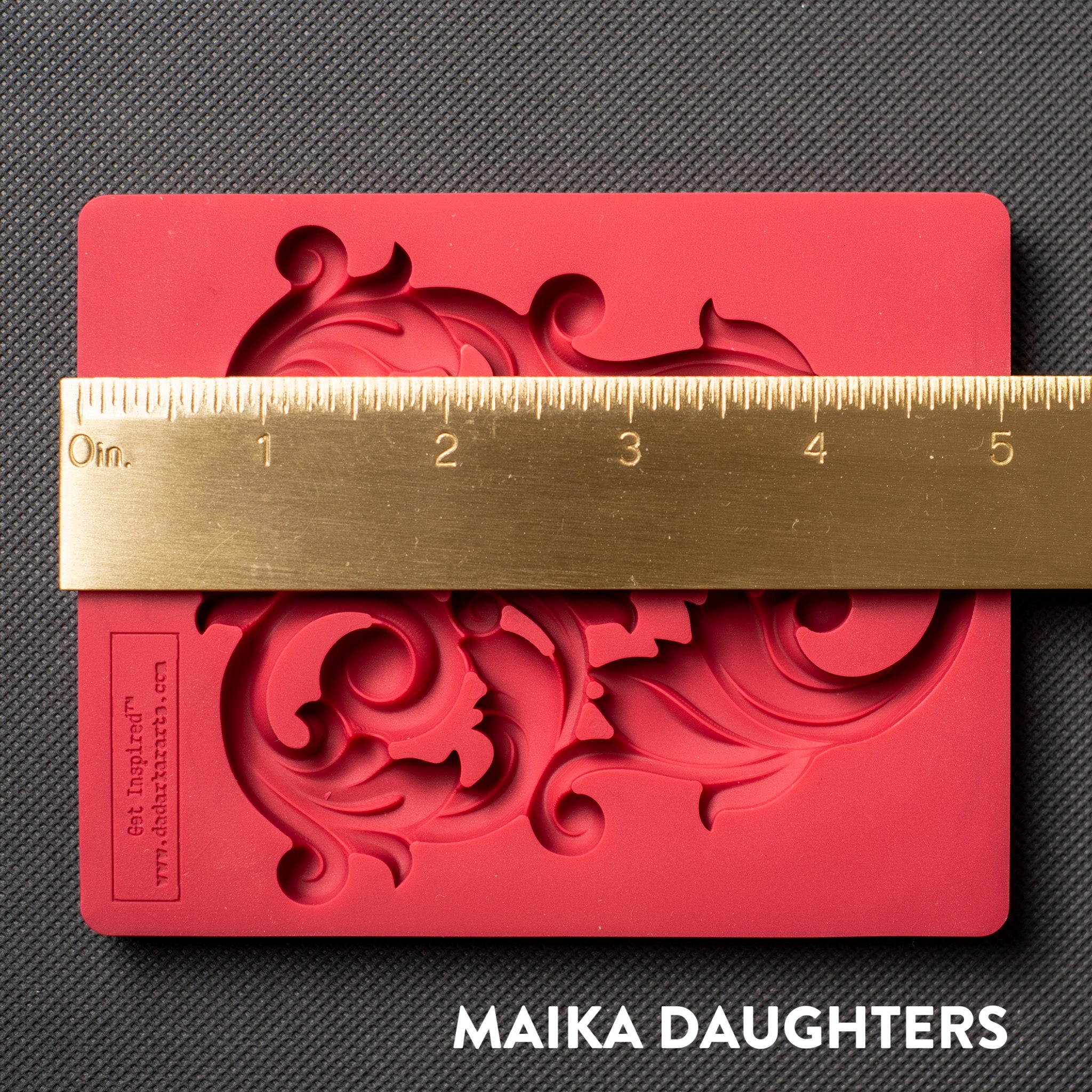 A red silicone mold that features an intricate flourish accent piece is against a dark grey material background. A gold ruler reading 5" length sits on the mold.