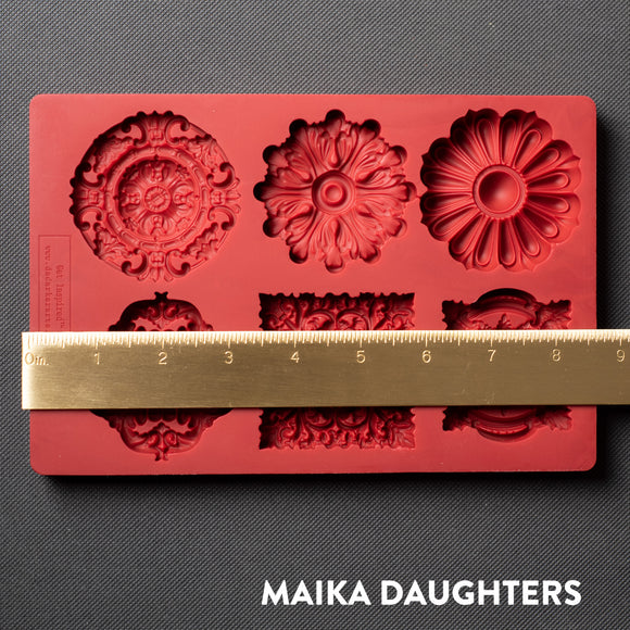 A red silicone mold featuring 6 ornate medallions is against a dark grey background. A gold ruler reading 8.5 inches width sits on the mold.