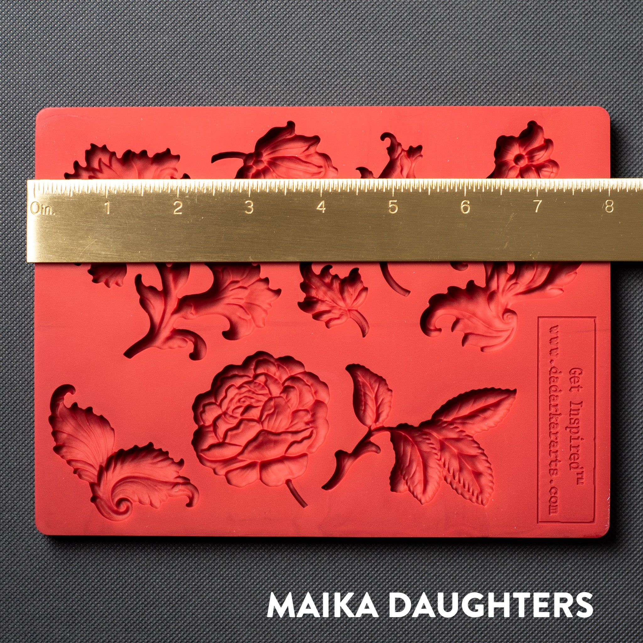 A red silicone mold featuring single flower blooms and loose leaves is against a dark grey material background. A gold ruler reading 8" length sits on the mold.