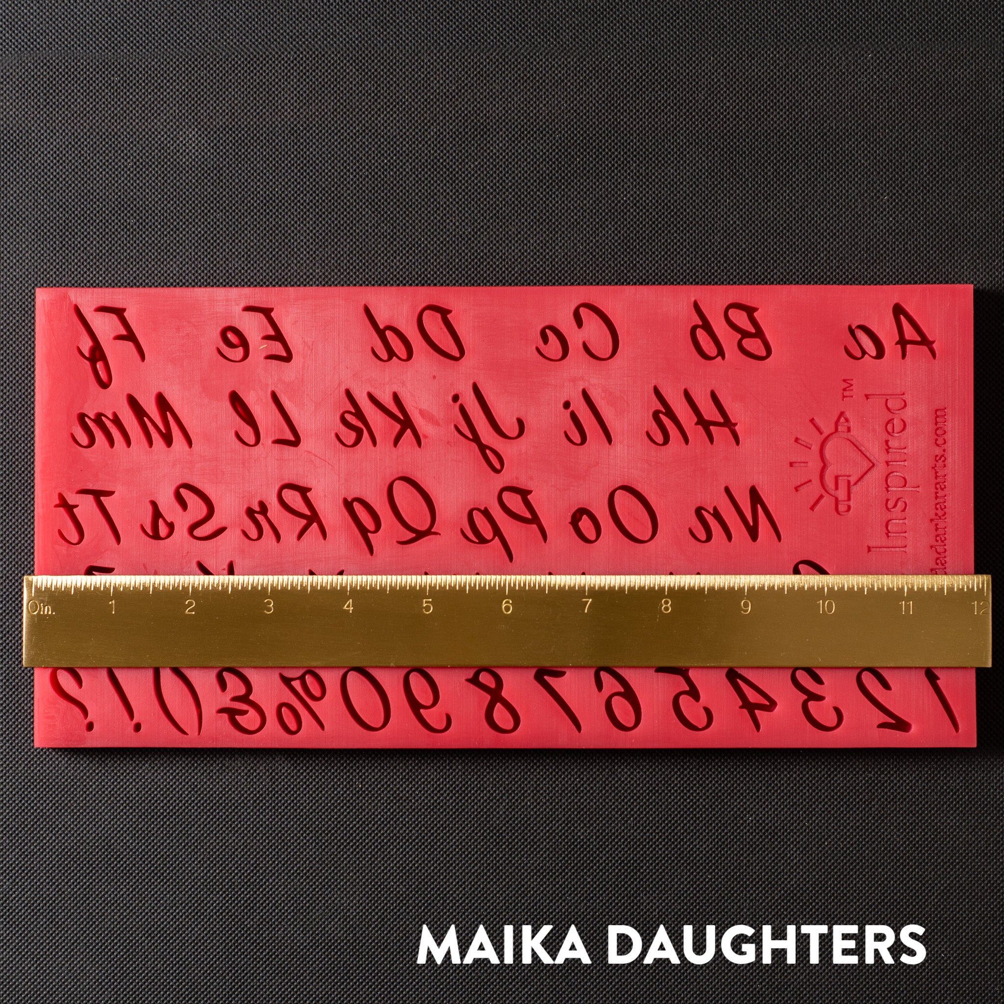 A red silicone mold of the English alphabet in script writing, numbers 0-9, and punctuations is against a dark grey background. A gold ruler reading 12" width sits on the mold.