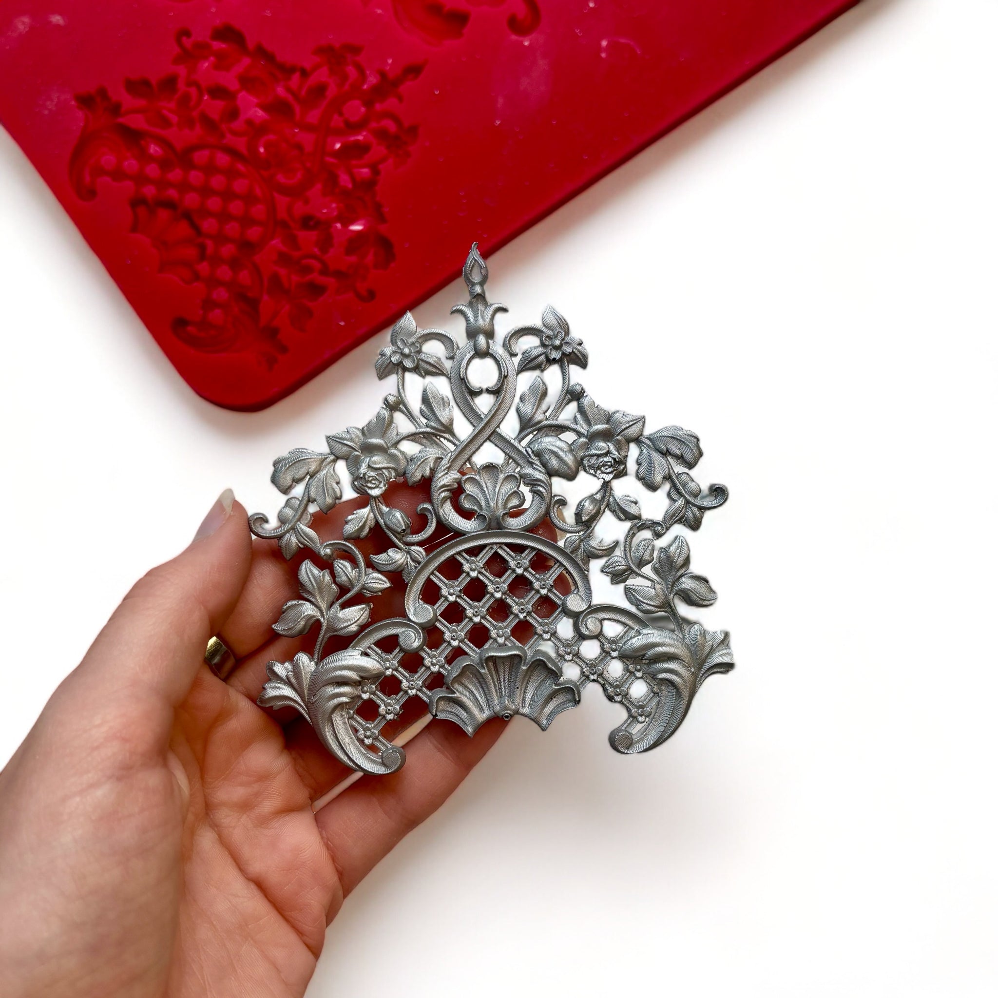 A red silicone mold and silver colored casting of 1 large vintage floral lattice center piece design are against a white background. A hand is shown holding the casting.