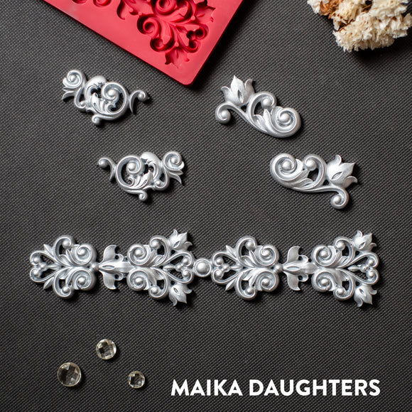 Silver colored silicone mold castings that features 4 small corner flourishes and one larger border are against a dark grey material background.