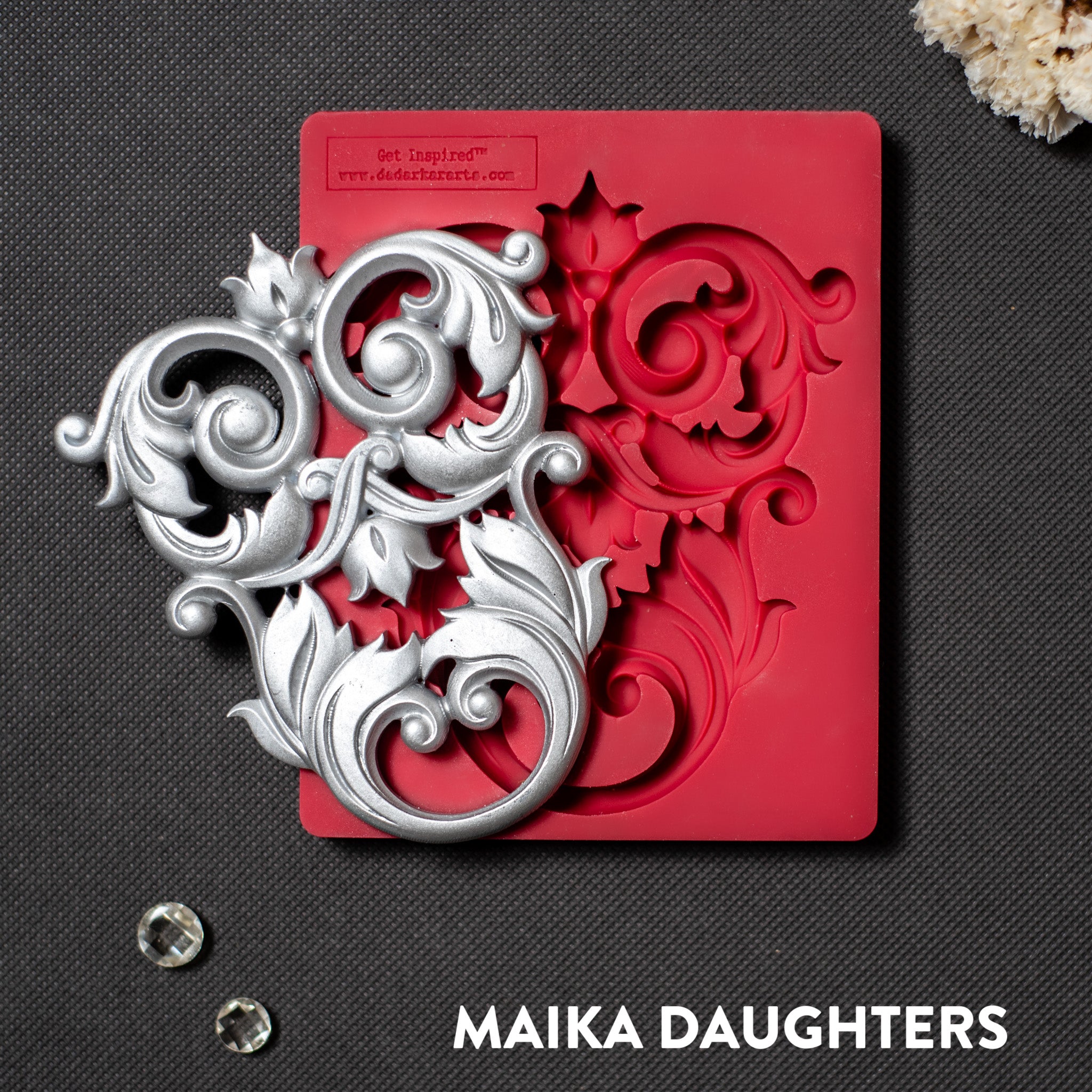 A red silicone mold and silver colored casting that features an intricate flourish accent piece is against a dark grey material background.