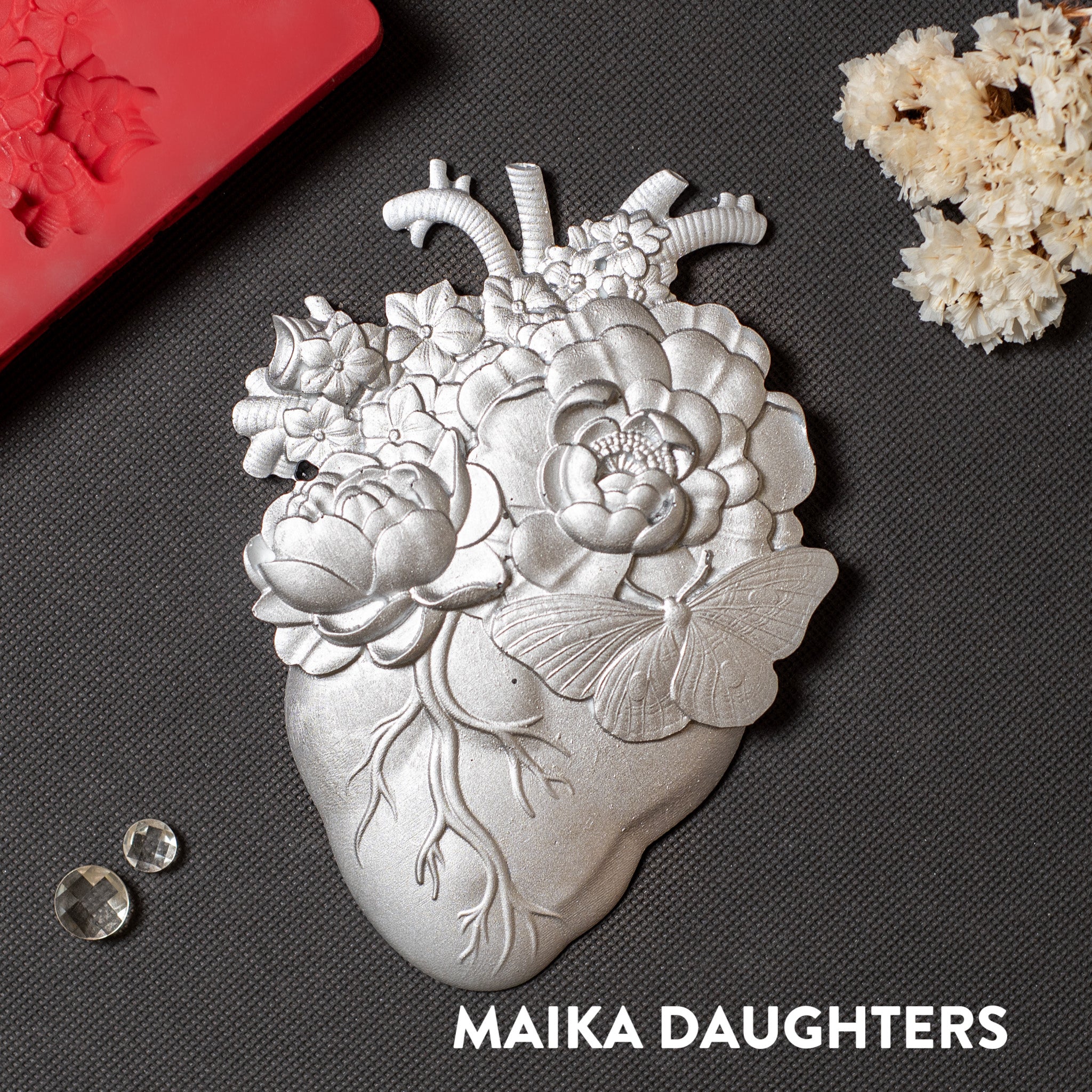 A silver colored silicone mold casting of an anatomical heart with flowers coming out of it is against a dark grey background.