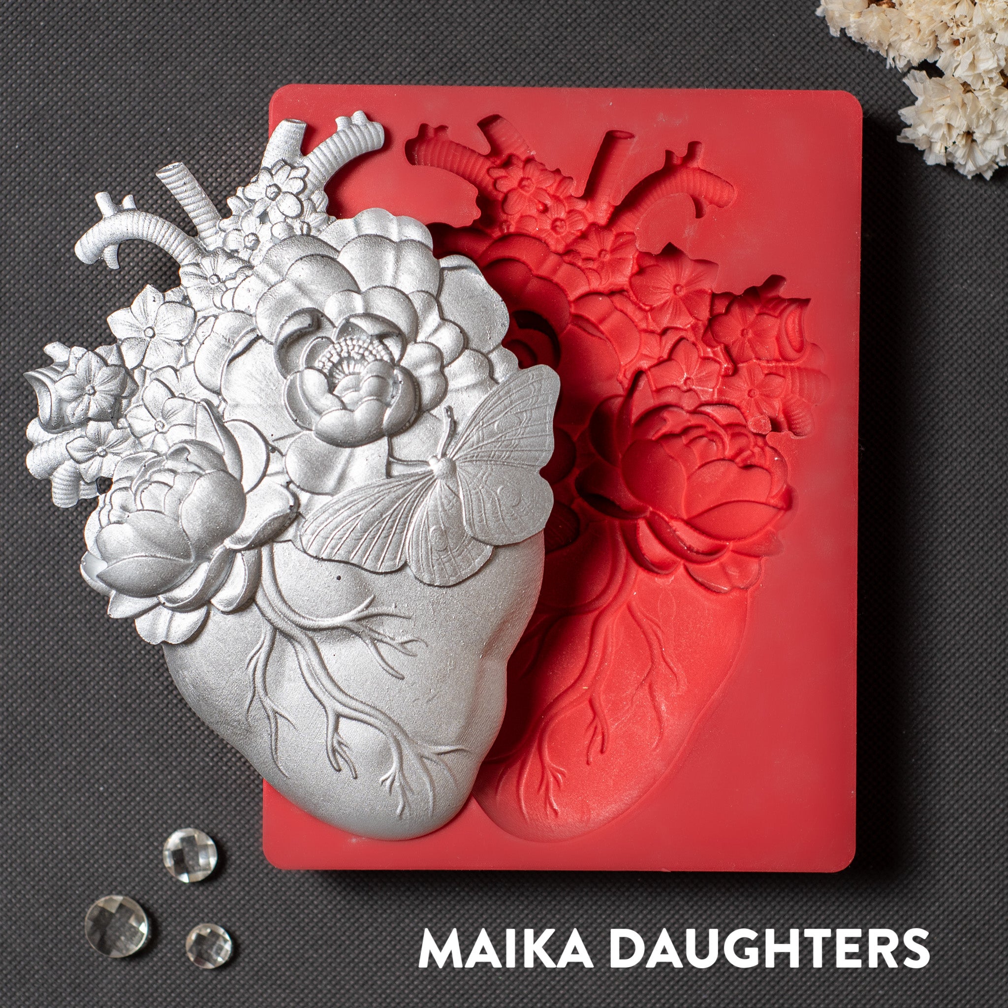 A red silicone mold and silver colored casting of an anatomical heart with flowers coming out of it are against a dark grey background.