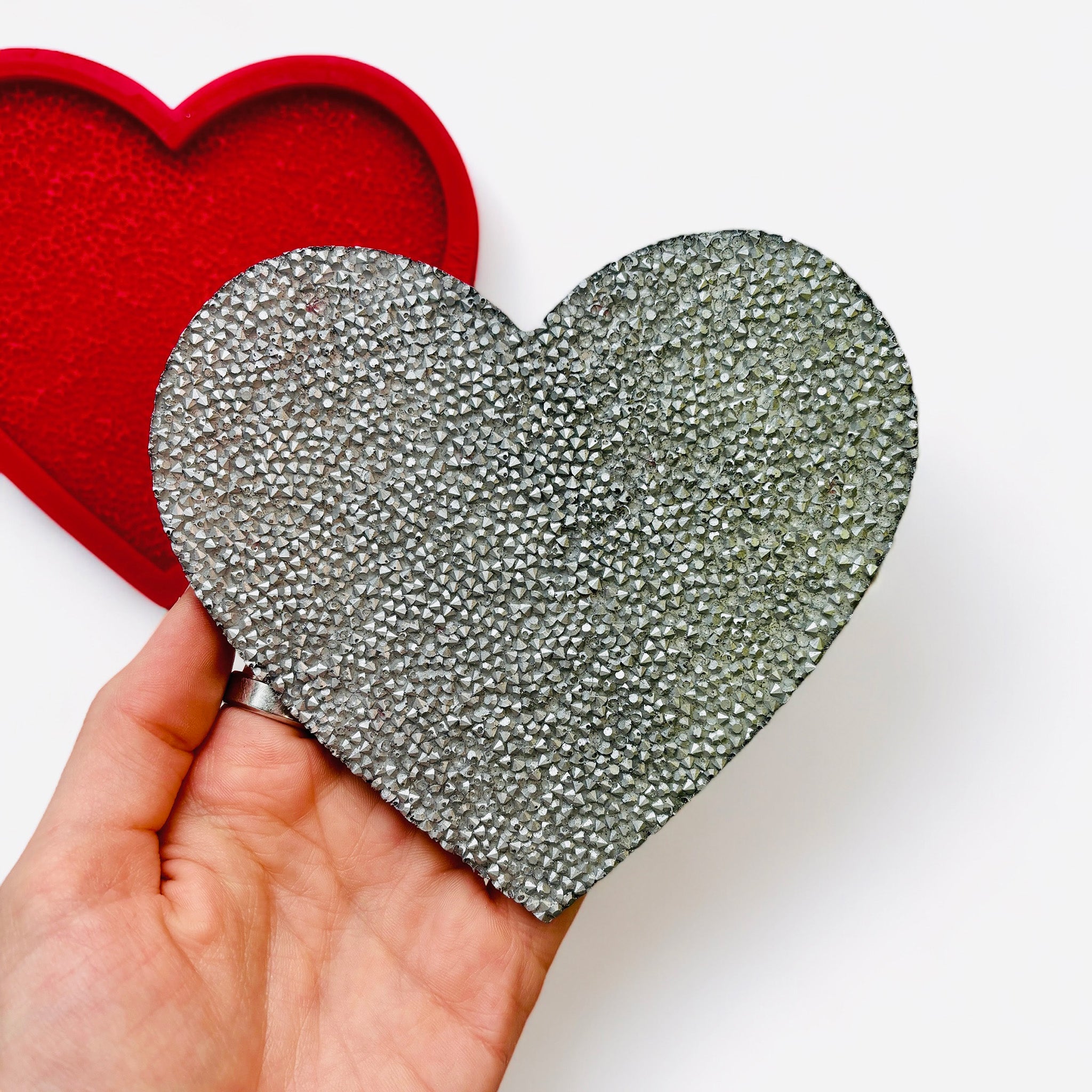 A red silicone mold and silver colored casting of a large textured heart are against a white background. A hand is shown holding the casting.