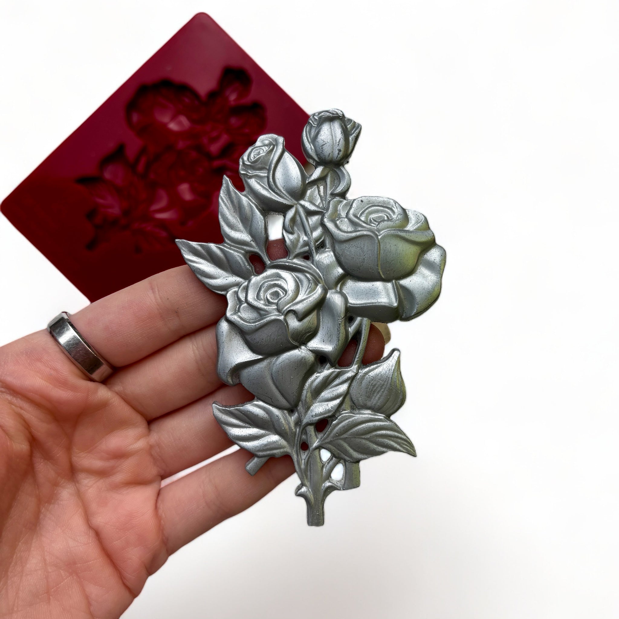 A red silicone mold and silver colored casting of a small bouquet of roses are against a white background. A hand is shown holding the casting.