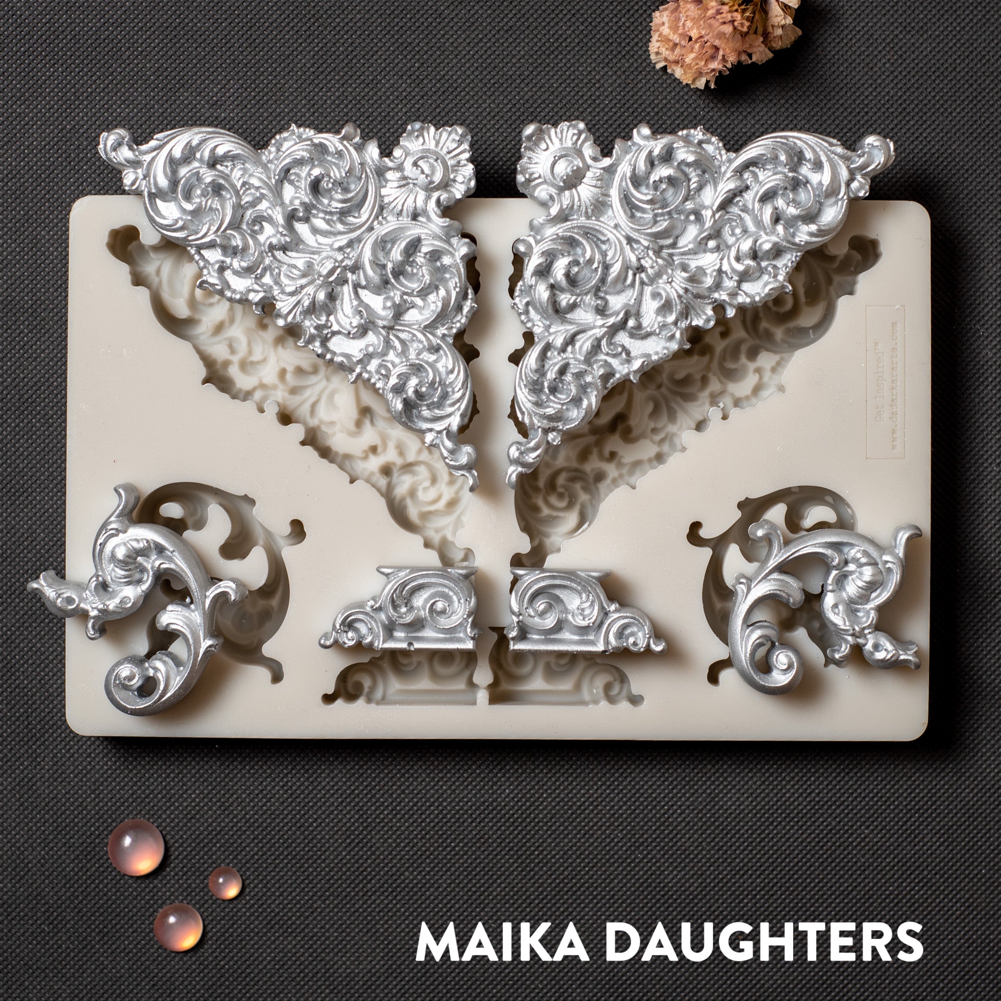 A light beige silicone mold and silver colored castings of ornate corner and accent pieces are against a dark grey background.