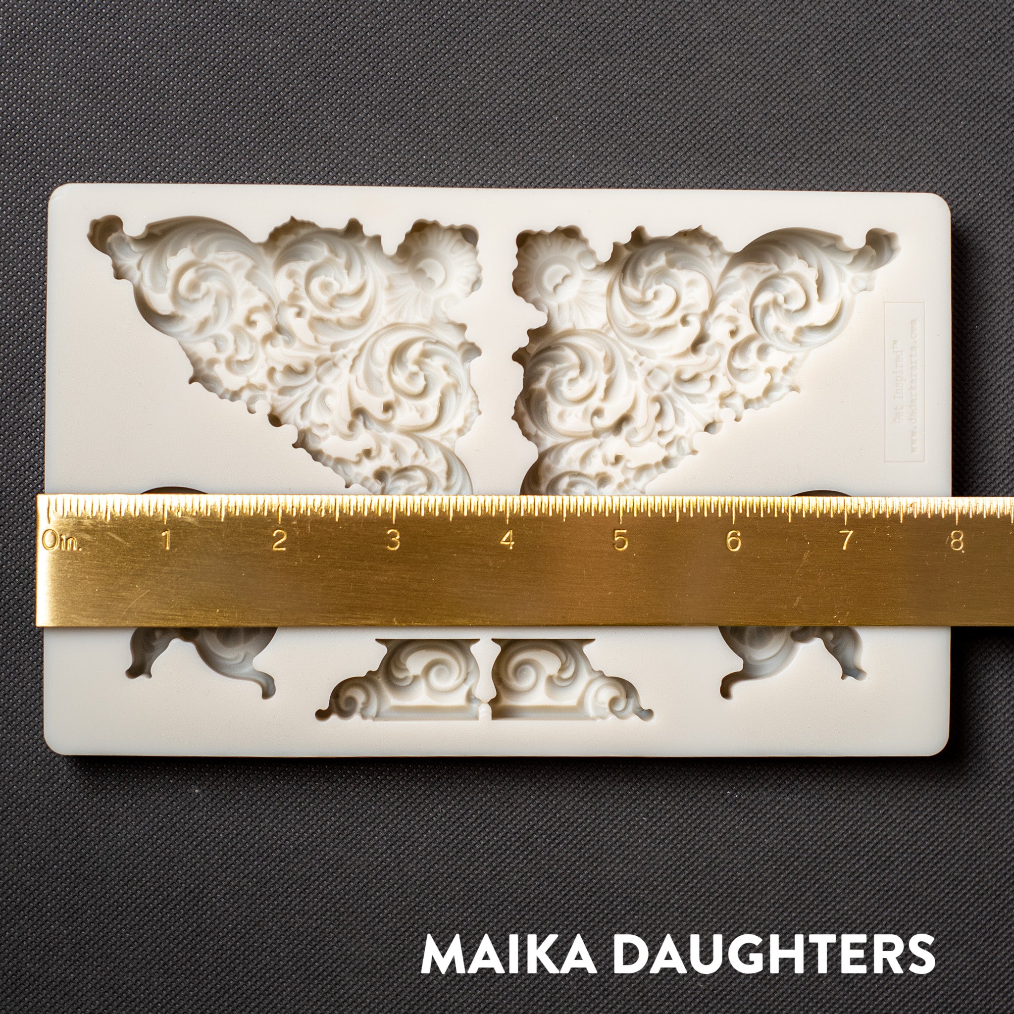 A light beige silicone mold of Get Inspired by Dadarkar Art's Illustrious Corners silicone mold is against a dark grey background. A gold ruler reading 8 inches width sits on the mold.