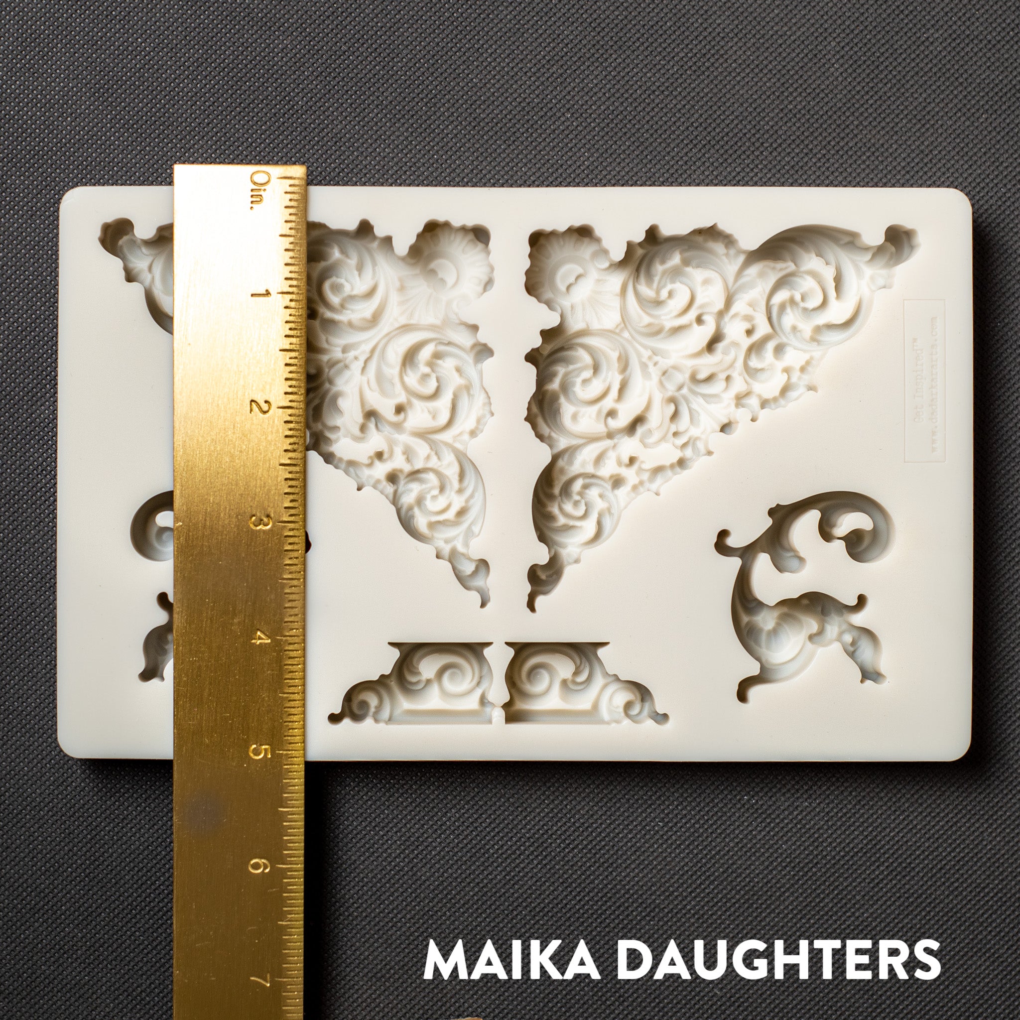 A light beige silicone mold of Get Inspired by Dadarkar Art's Illustrious Corners silicone mold is against a dark grey background. A gold ruler reading 5.25 inches height sits on the mold.