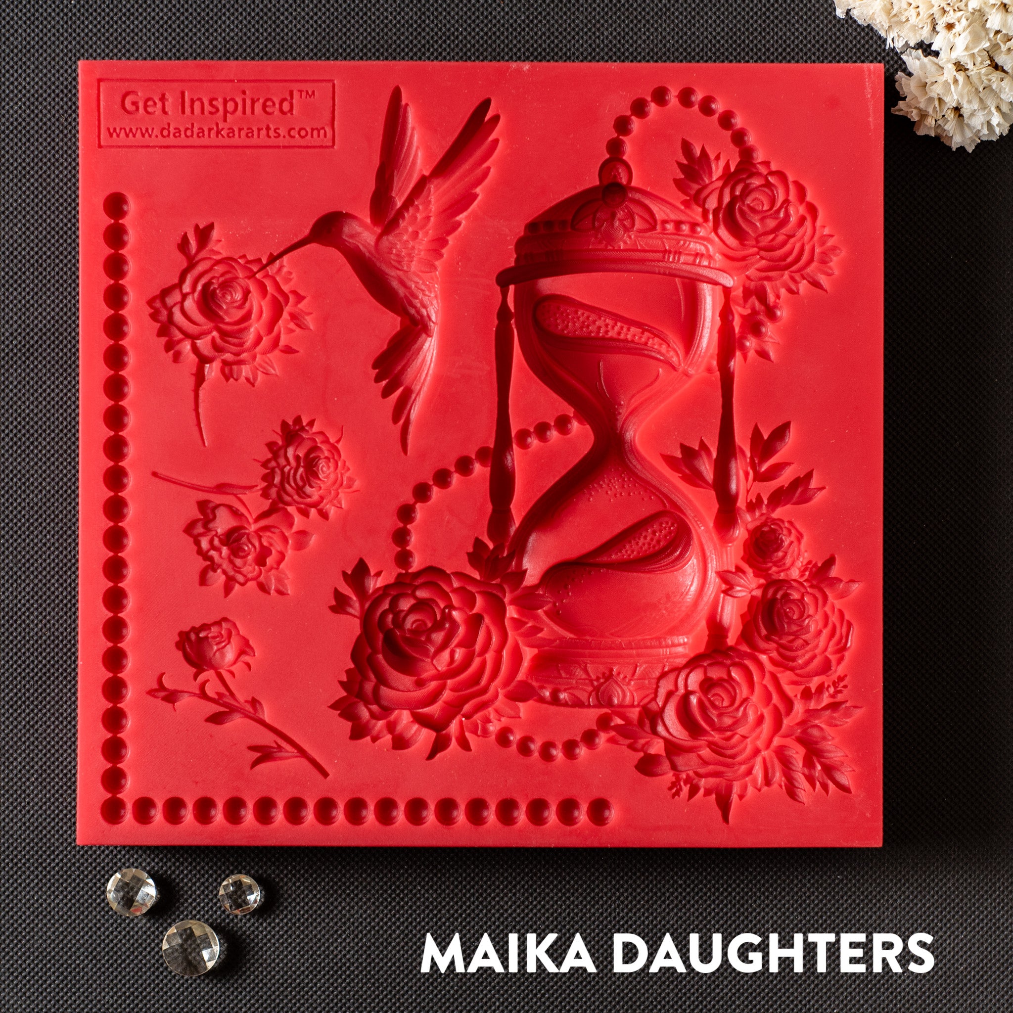 A red silicone mold featuring hourglass wrapped in intricate flowers and beads, a hummingbird, flower blooms, and a beaded corner trim is against a dark grey background.