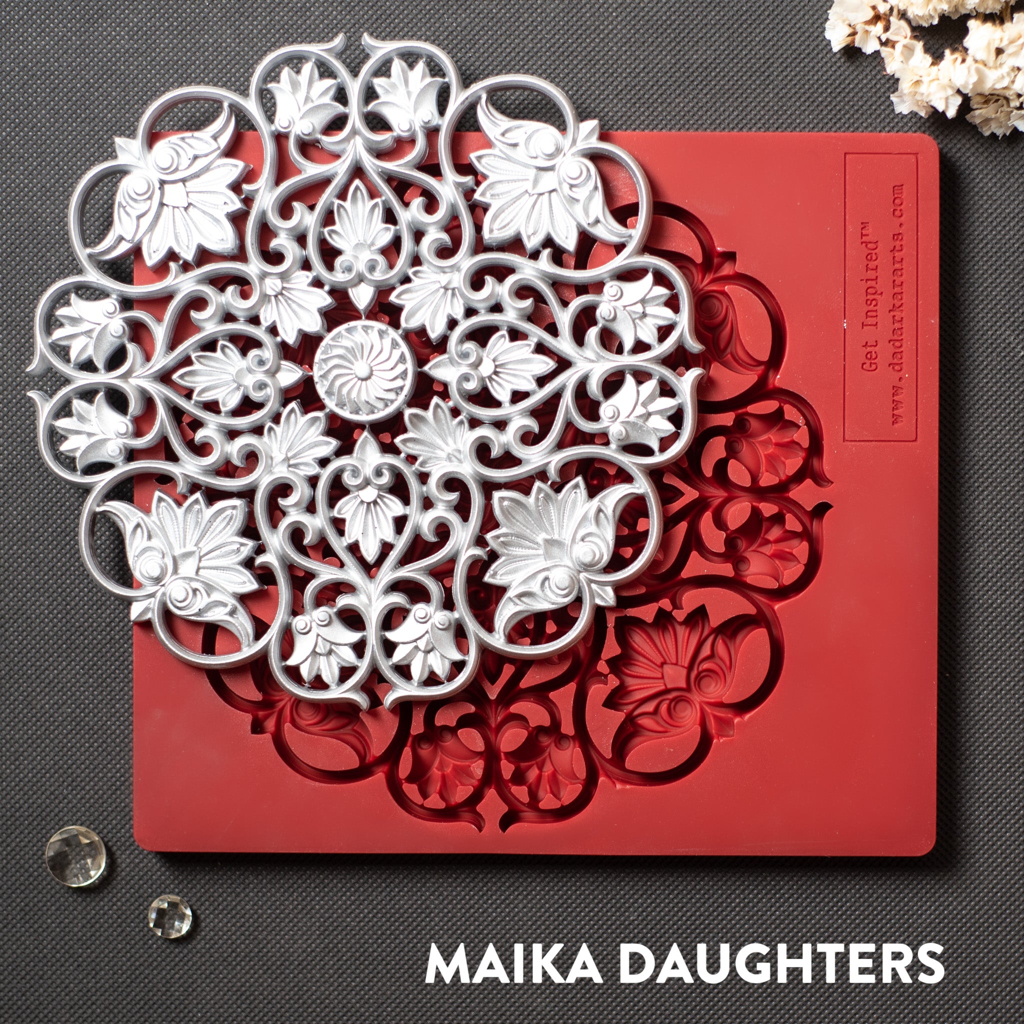 A red silicone mold and a silver colored casting of a large mandala with lotus flowers in it are against a dark grey background.