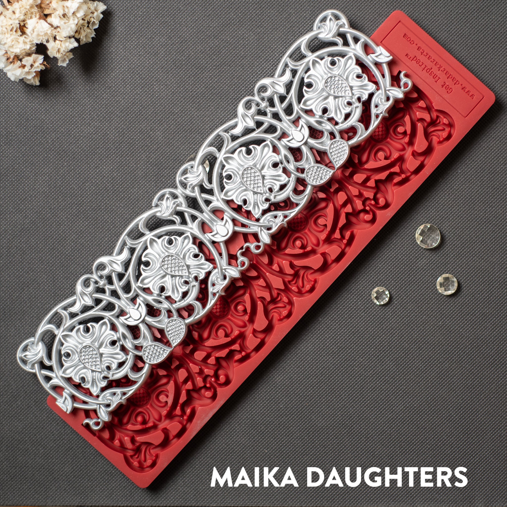 A red silicone mold and silver colored casting of an ornate border is against a dark grey background.