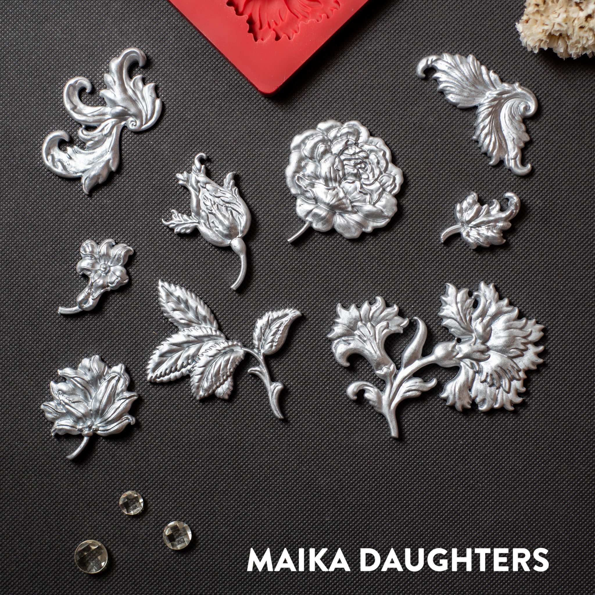 Silver colored silicone mold castings featuring single flower blooms and loose leaves are against a dark grey material background.