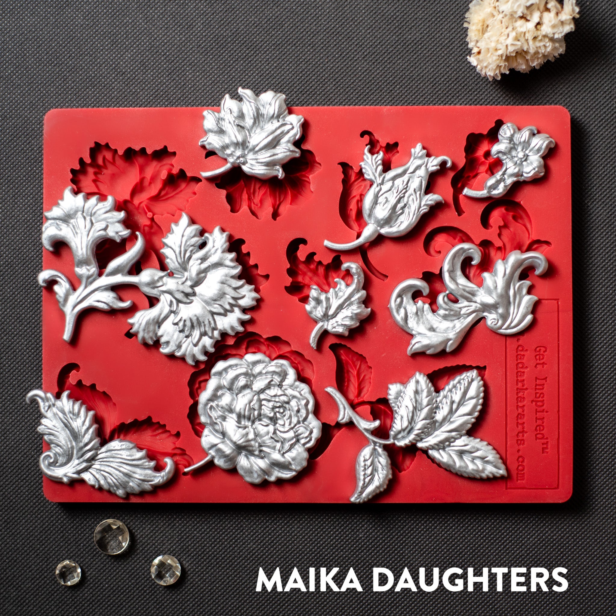 A red silicone mold and silver colored casting featuring single flower blooms and loose leaves are against a dark grey material background.