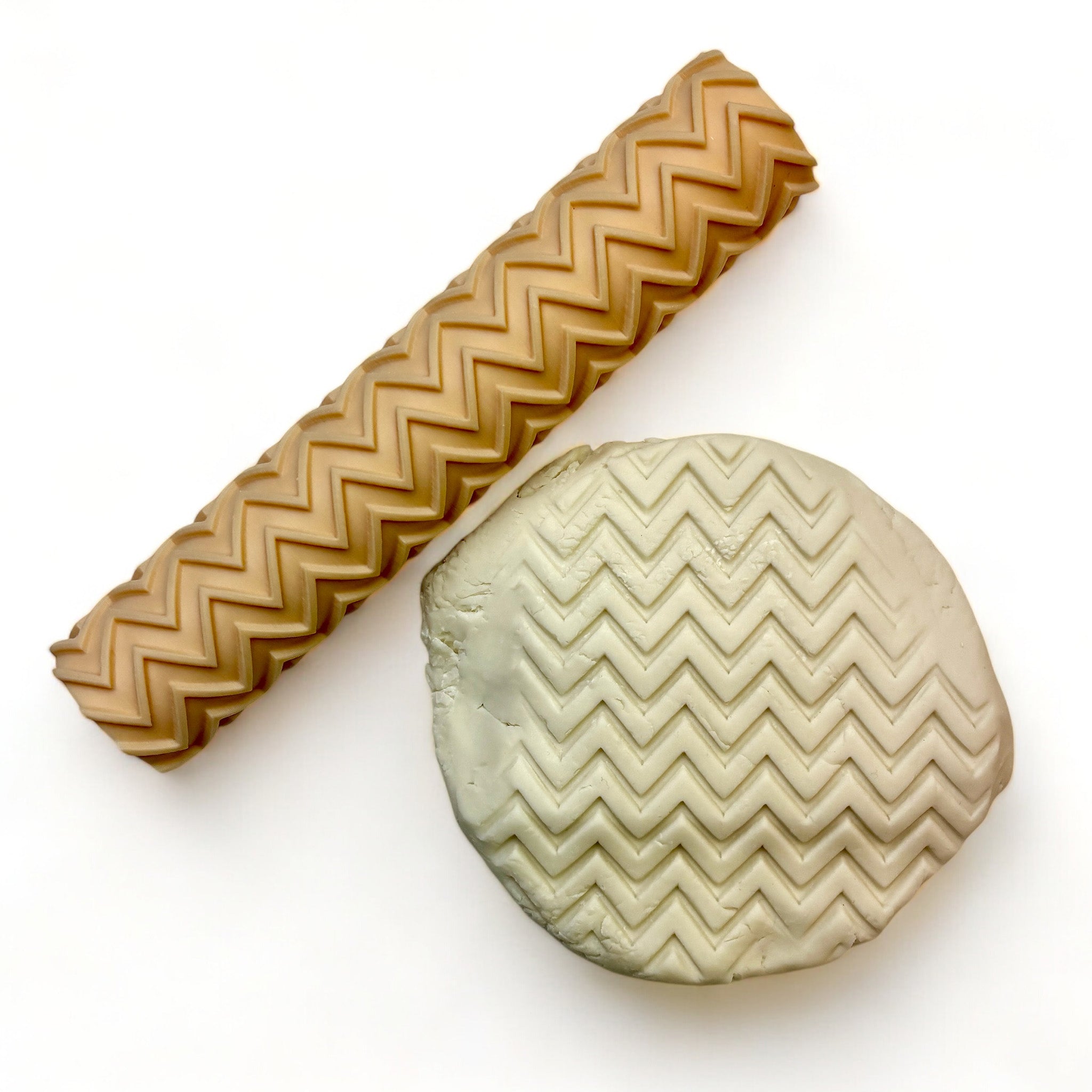 A seamless texture roller and flattened chunk of clay featuring a zig zag pattern are against a white background.
