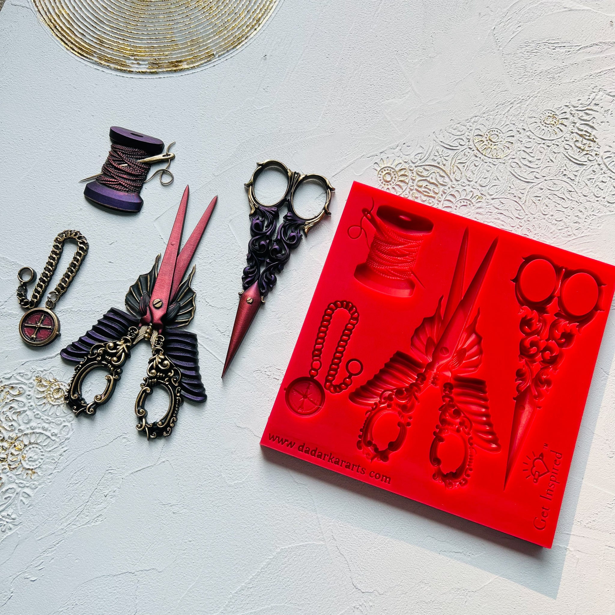 A red silicone mold and colorful castings of 2 steampunk style sewing scissors, a spool of thread with a needle in it, and a small pocket compass are against a white background.